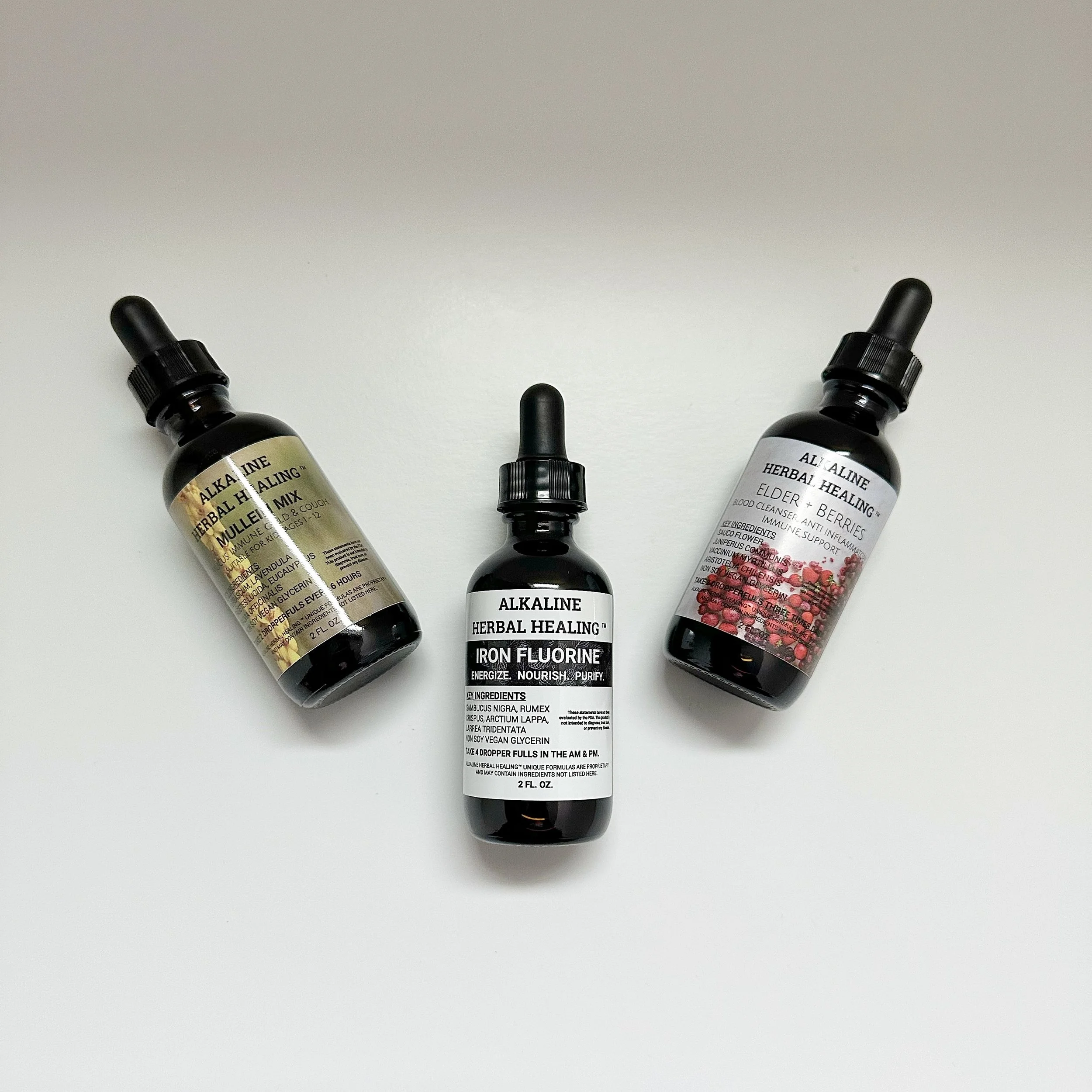 Three black dropper bottles labeled 'Alkaline Herbal Healing' with herbal and berry images on the labels, arranged on a white surface.