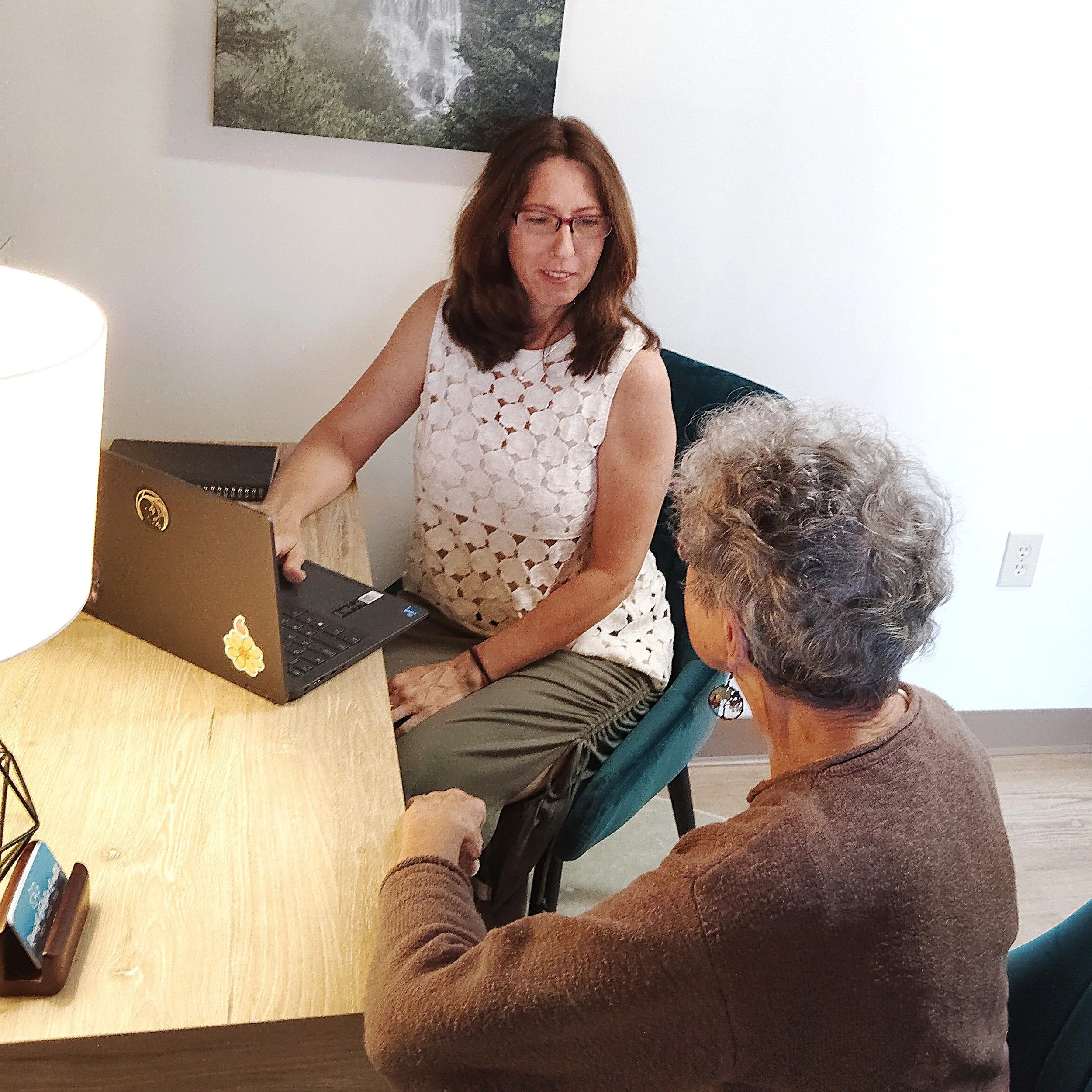 A woman with glasses in a white dress with floral holes using a laptop at a wooden table, surrounded by three older adults, with a painting of a waterfall in a forest on the wall.