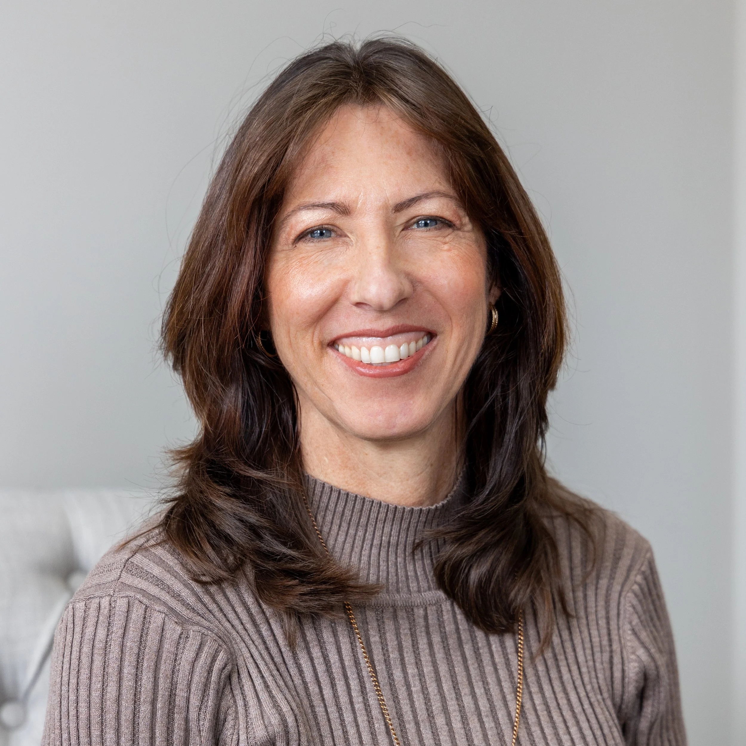 A smiling woman with long brown hair and blue eyes, wearing a beige ribbed sweater and a gold chain necklace, standing indoors against a plain gray background.