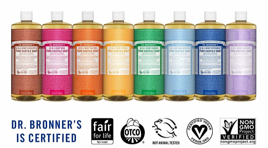 A row of eight bottles of Dr. Bronner's Pure-Castile Soap in different scents, with labels in various colors. Beneath the bottles are certification icons, including fair trade, OTC, not animal tested, vegan, and non-GMO.