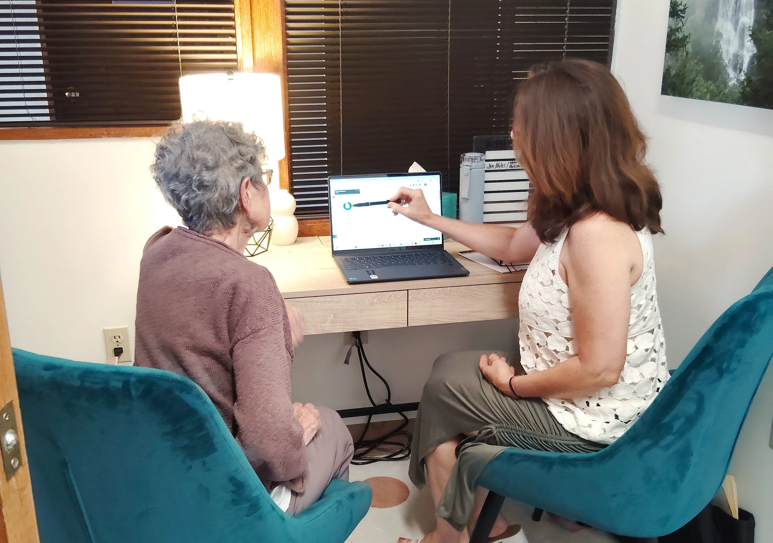 A woman showing an elderly woman a computer screen while sitting in a room with wooden blinds, a lamp, and a framed picture of a river or waterfall on the wall.