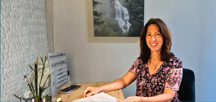 A woman with shoulder-length brown hair, wearing a floral patterned shirt, sitting at a desk with papers, smiling, with a waterfall picture on the wall behind her.