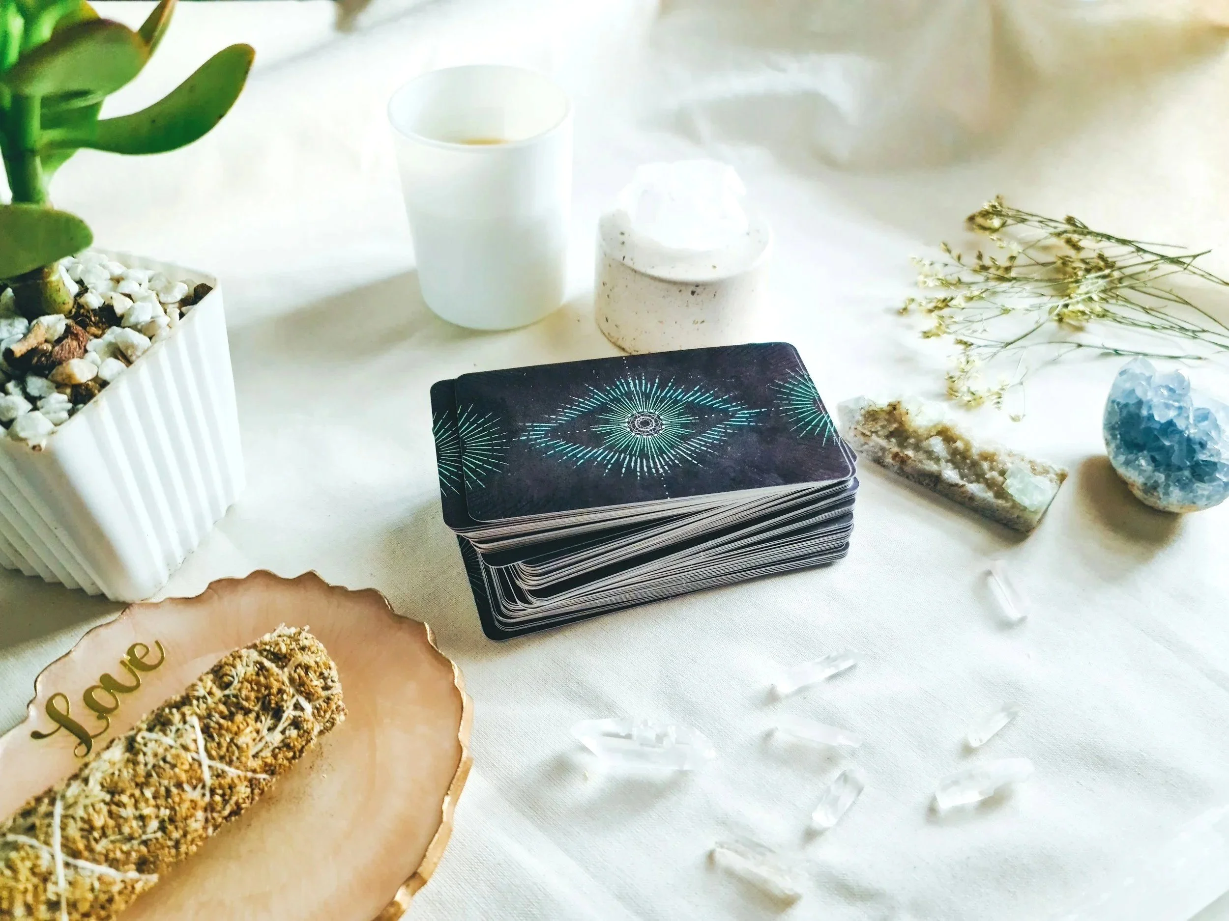 A table with a potted plant, a white cup with a beverage, a white container, a deck of tarot cards, crystals, and a plate of cookies with the word "love" written on it.