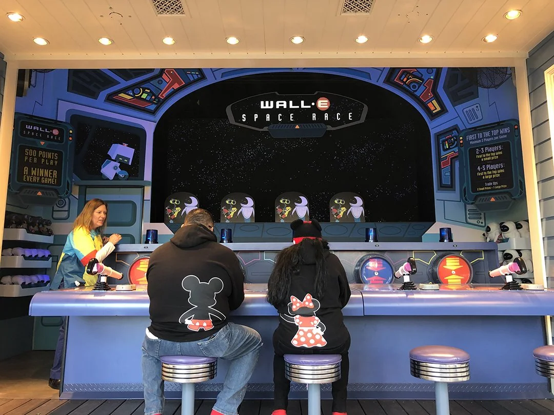 People playing a space-themed arcade game called 'Wall-E Space Race' in an indoor arcade, with a space-themed backdrop and game instructions displayed on screens.
