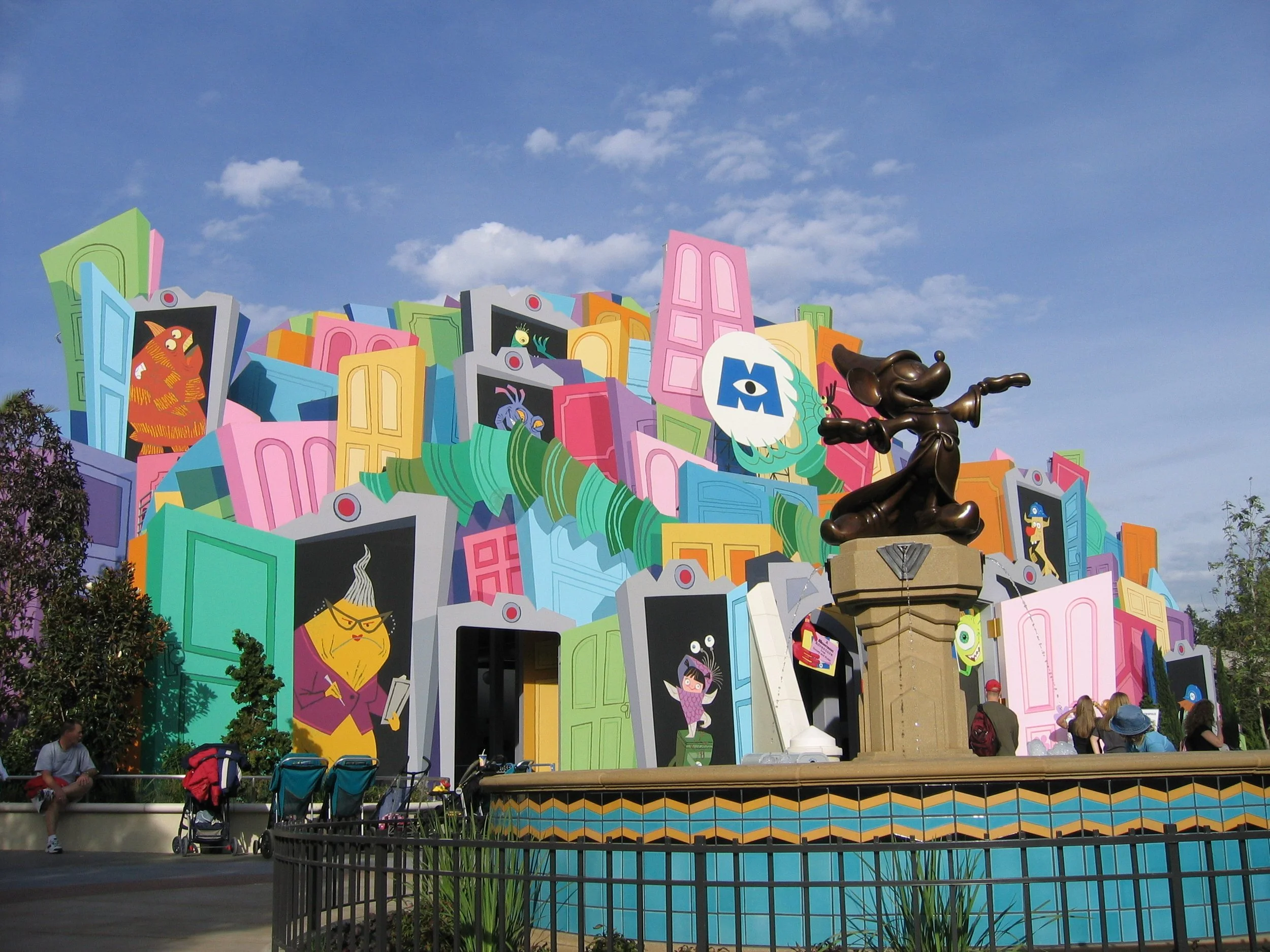 Colorful mural with cartoon characters and doors, with a bronze character statue in the foreground, and people sitting and standing around, under a bright blue sky with scattered clouds.