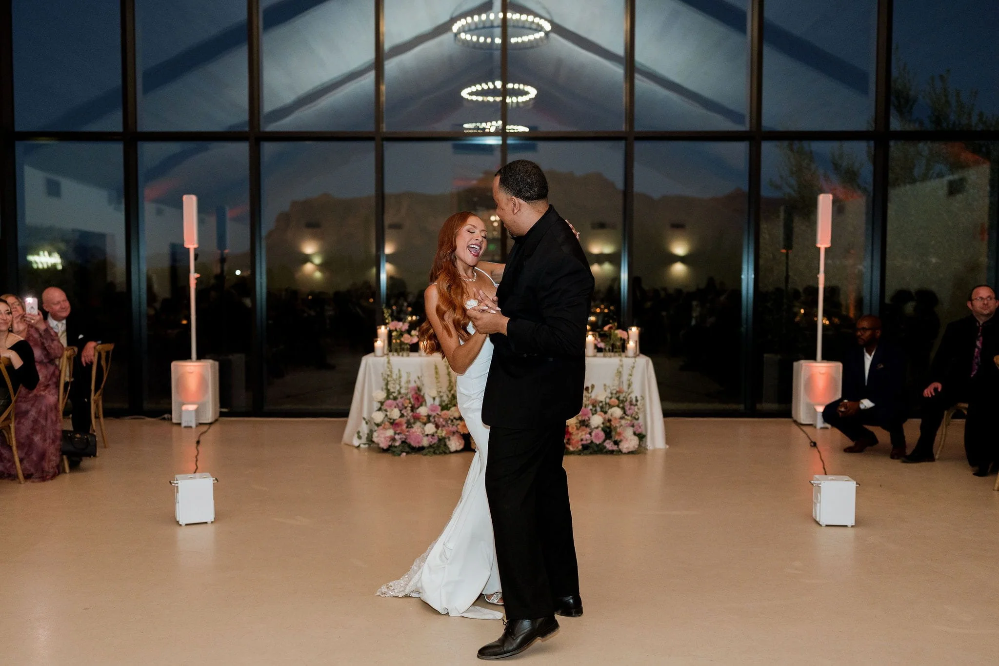 A couple dancing at a wedding reception, surrounded by guests and floral arrangements.