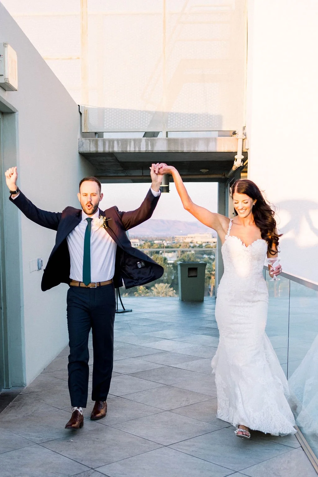 A couple joyfully walking on a rooftop patio, with the man in a dark suit and the woman in a white lace gown, holding hands in celebration.