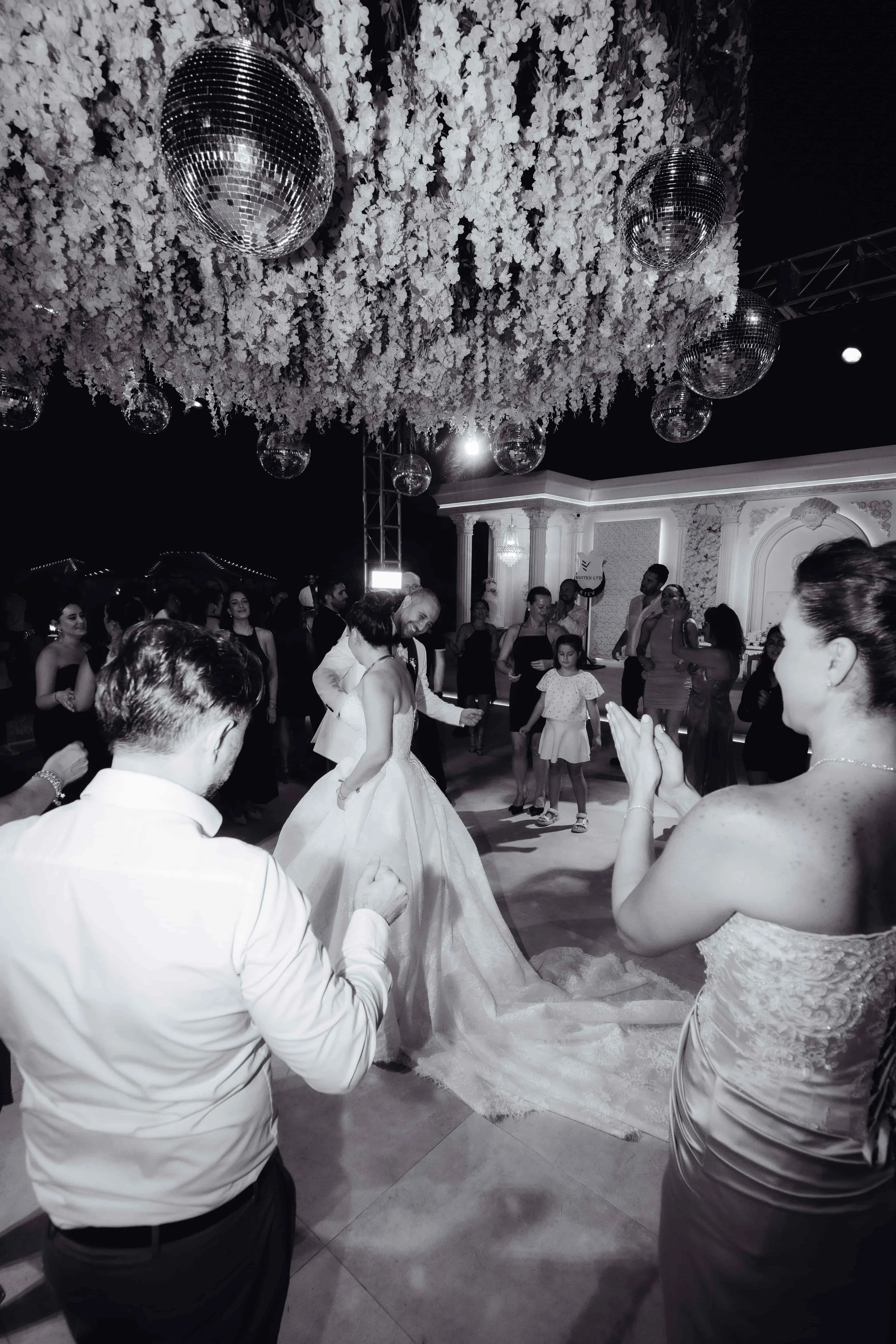 Black-and-white wedding reception photo showing a bride dancing with a man beneath hanging flowers and mirrored disco balls while guests watch and clap.