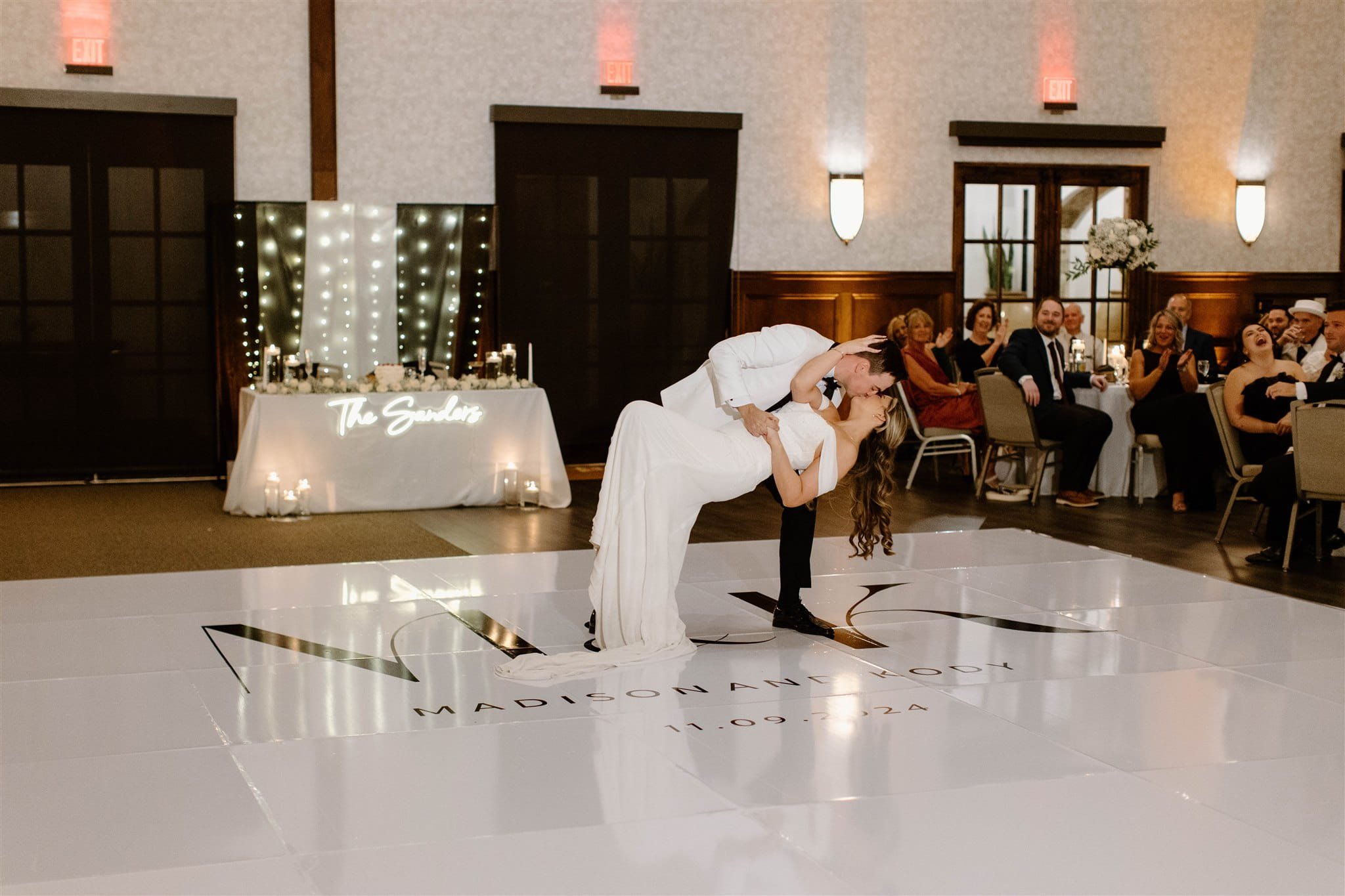 Newlywed couple kisses while dancing on a decorated reception floor as seated guests watch.