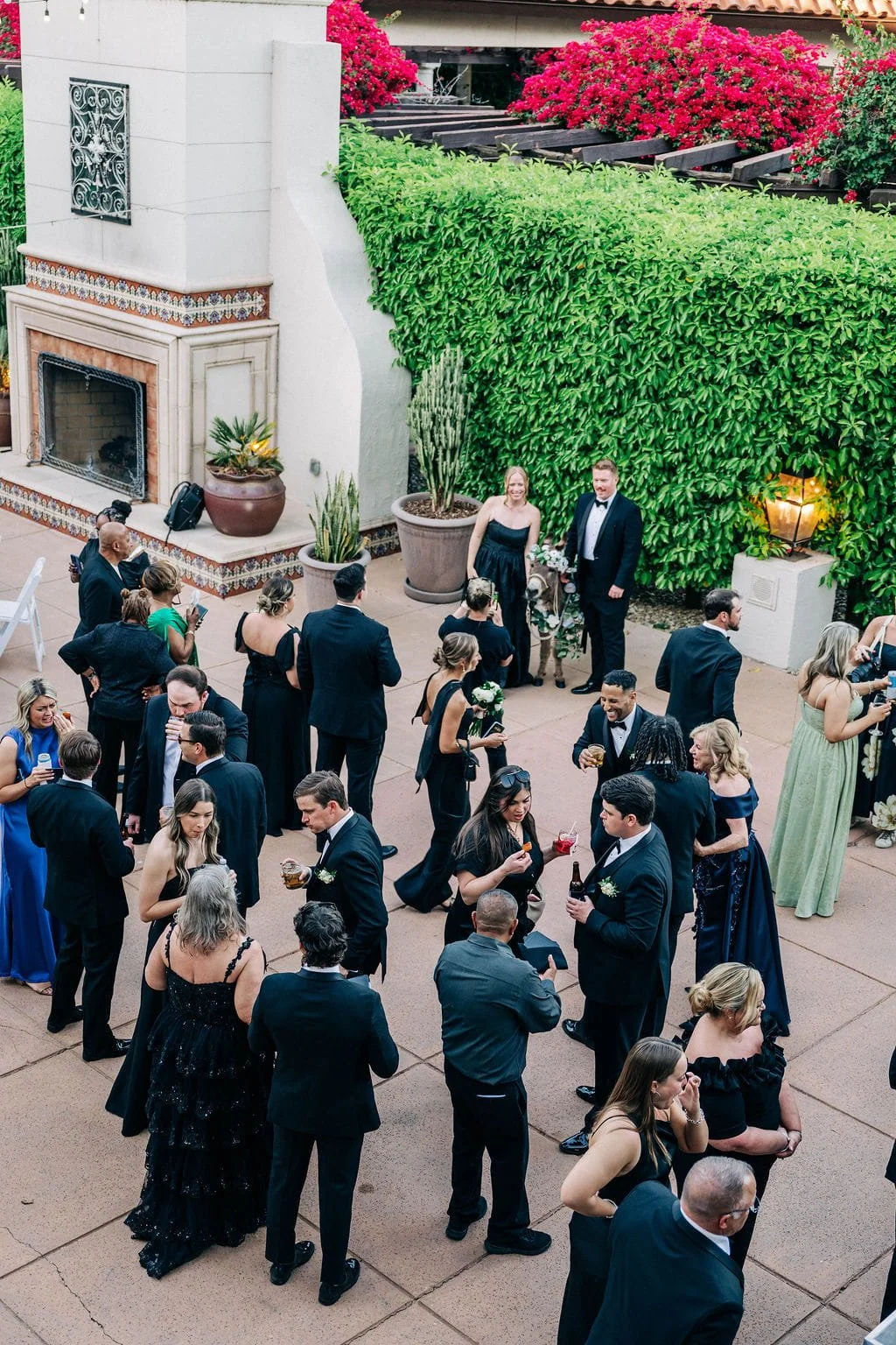 Formally dressed guests mingle at an outdoor wedding reception in a landscaped courtyard with a tiled fireplace, flowering bushes, and a tall green hedge.