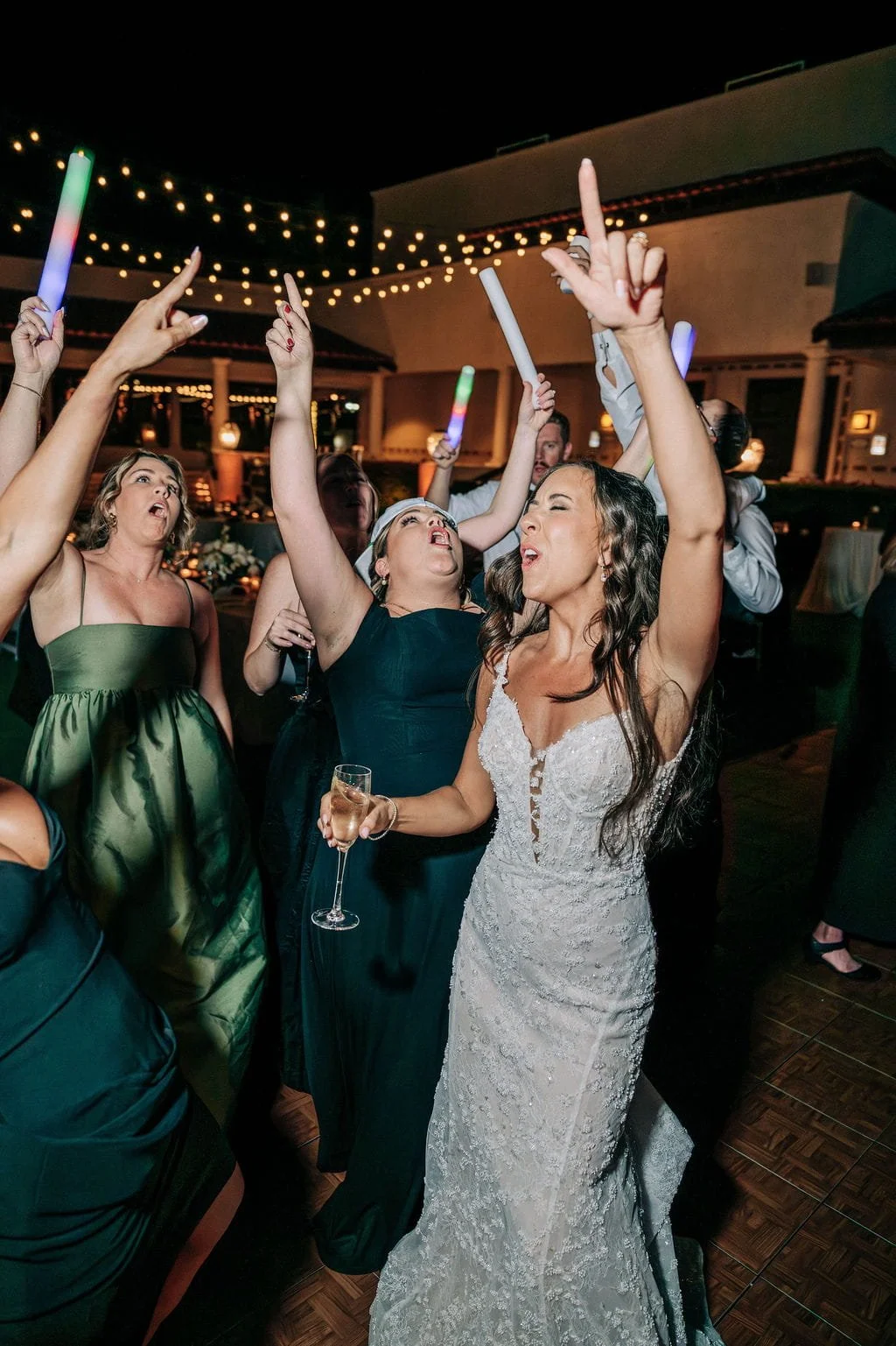 A lively party scene with a woman in a white lace wedding dress holding a champagne glass, surrounded by people dancing with colorful light sticks under string lights.