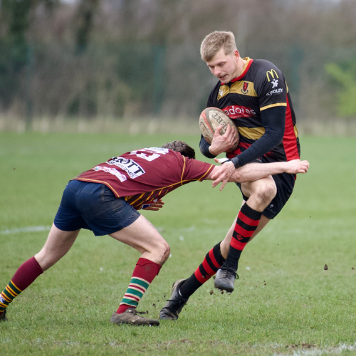 Rugby players in black, red, and yellow uniforms playing on a grassy field with trees in the background, one player is tackling another while holding a rugby ball.