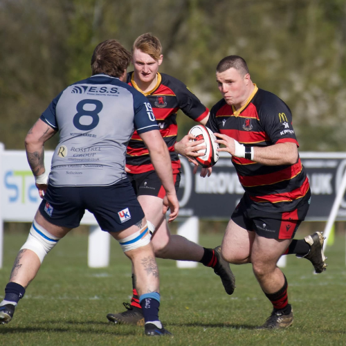 A rugby player in a black and red uniform running on the field holding a rugby ball while spectators watch from the sidelines.