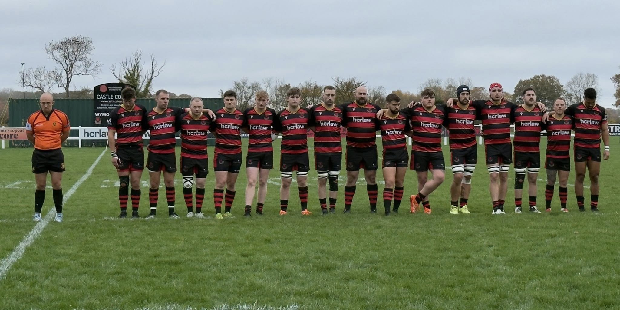 Rugby team standing in a line on a field with a referee on the left, wearing black and red striped jerseys and black shorts, with trees and a signpost in the background.