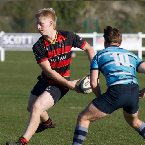 A rugby game with players on the field. One player in black and red is running with the ball, while three players in blue and red are on the ground in the background, one on his stomach and two on their knees.