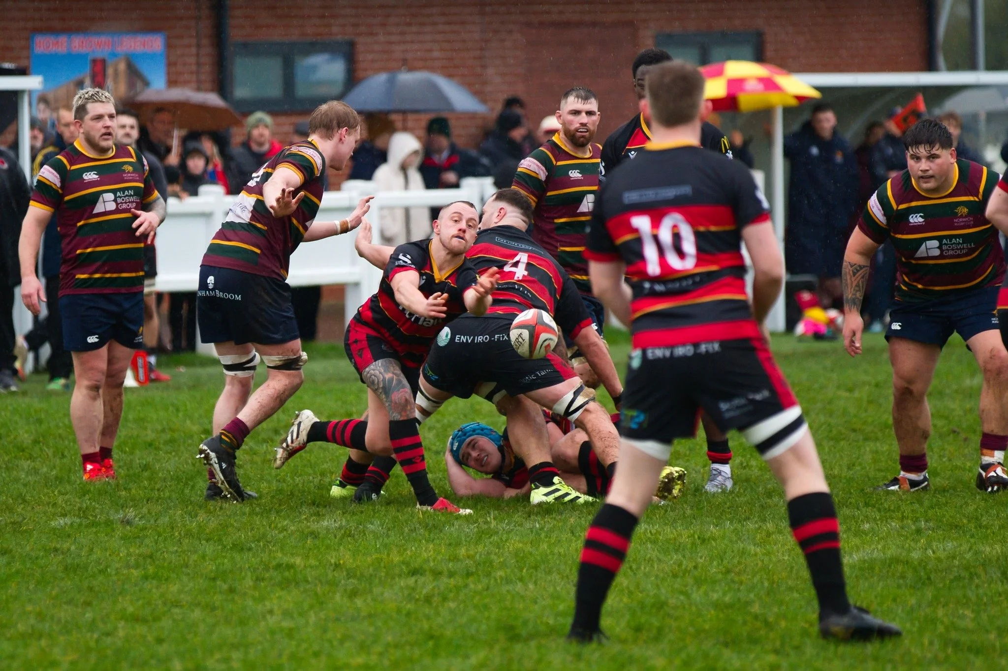 Rugby players in black and red stripes engaged in a tackle during a match, with others observing on a green field, rainy weather, and some spectators with umbrellas in background.