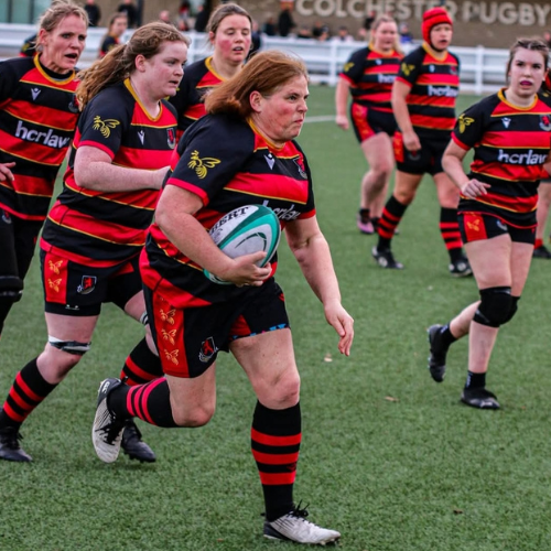 Two female rugby players in black, red, and gold uniforms on the field. One is running with a rugby ball, and the other is running beside her. There are other players and a coach or spectator in the background.