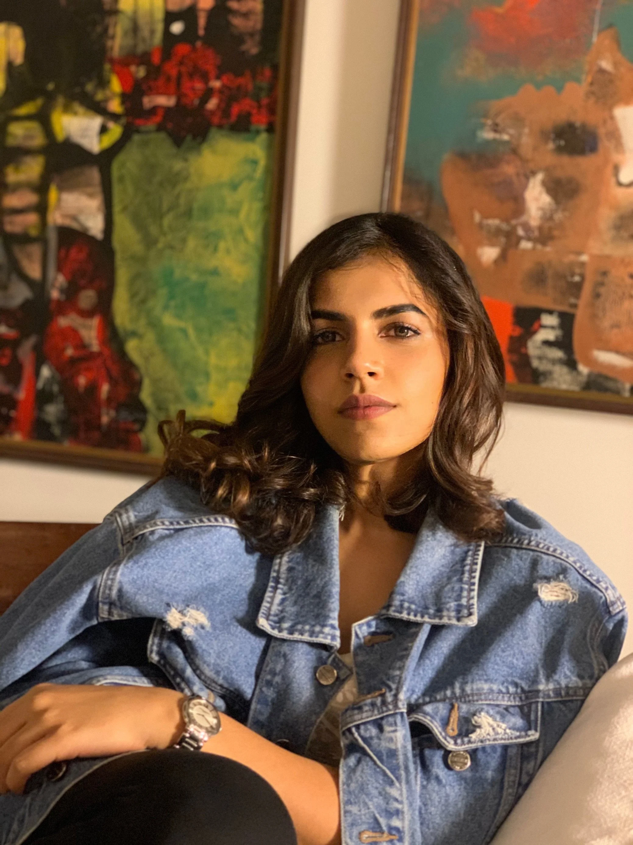 A young woman with brown, wavy hair wearing a denim jacket and a watch, sitting at a table with abstract paintings on the wall behind her.