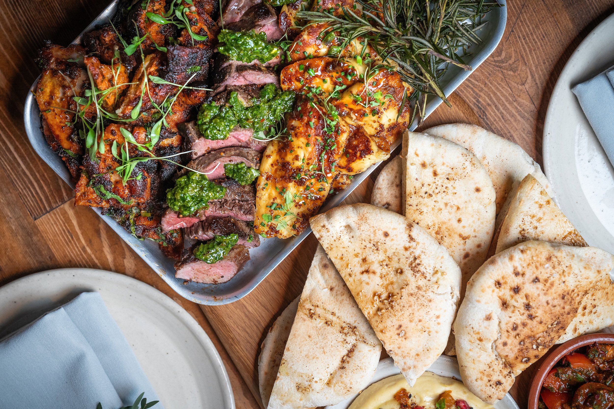 A tray of sliced fillet steak garnished with herbs, grilled chicken with sauce, and fresh herbs on a metal platter. Nearby, there are pieces of pita bread, a bowl of hummus with toppings, and a side of vegetable salad.