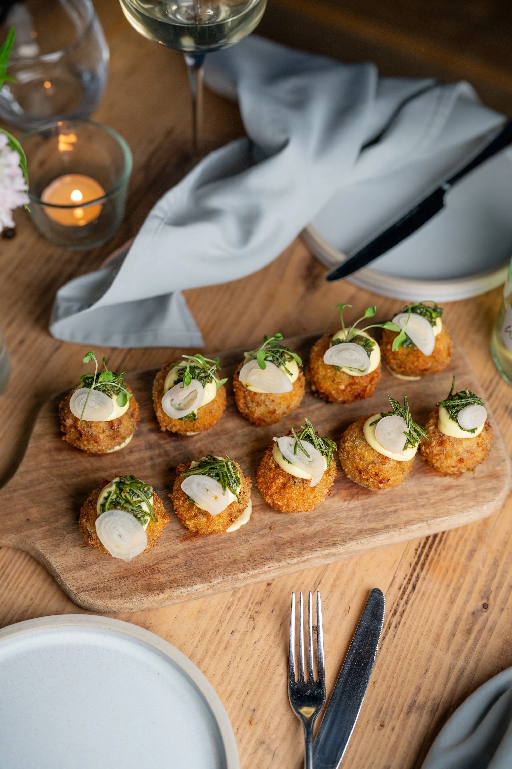 A plated gourmet dish with a base of squash puree, topped with green herbs, scallops, and slices of crispy pancetta. In the background, there is a glass of water with greenery and another plate with a layered dish on a wooden table.