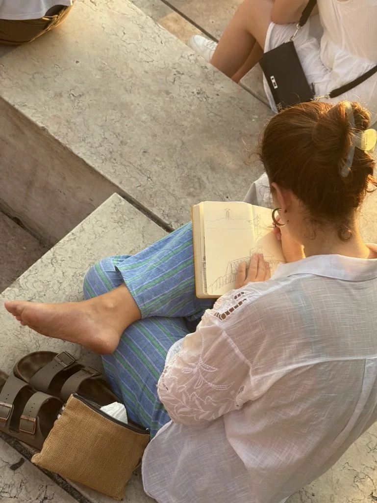 Una mujer dibujando en un cuaderno sentado en un banco de piedra mientras otra persona está sentada cerca, en un lugar al aire libre.