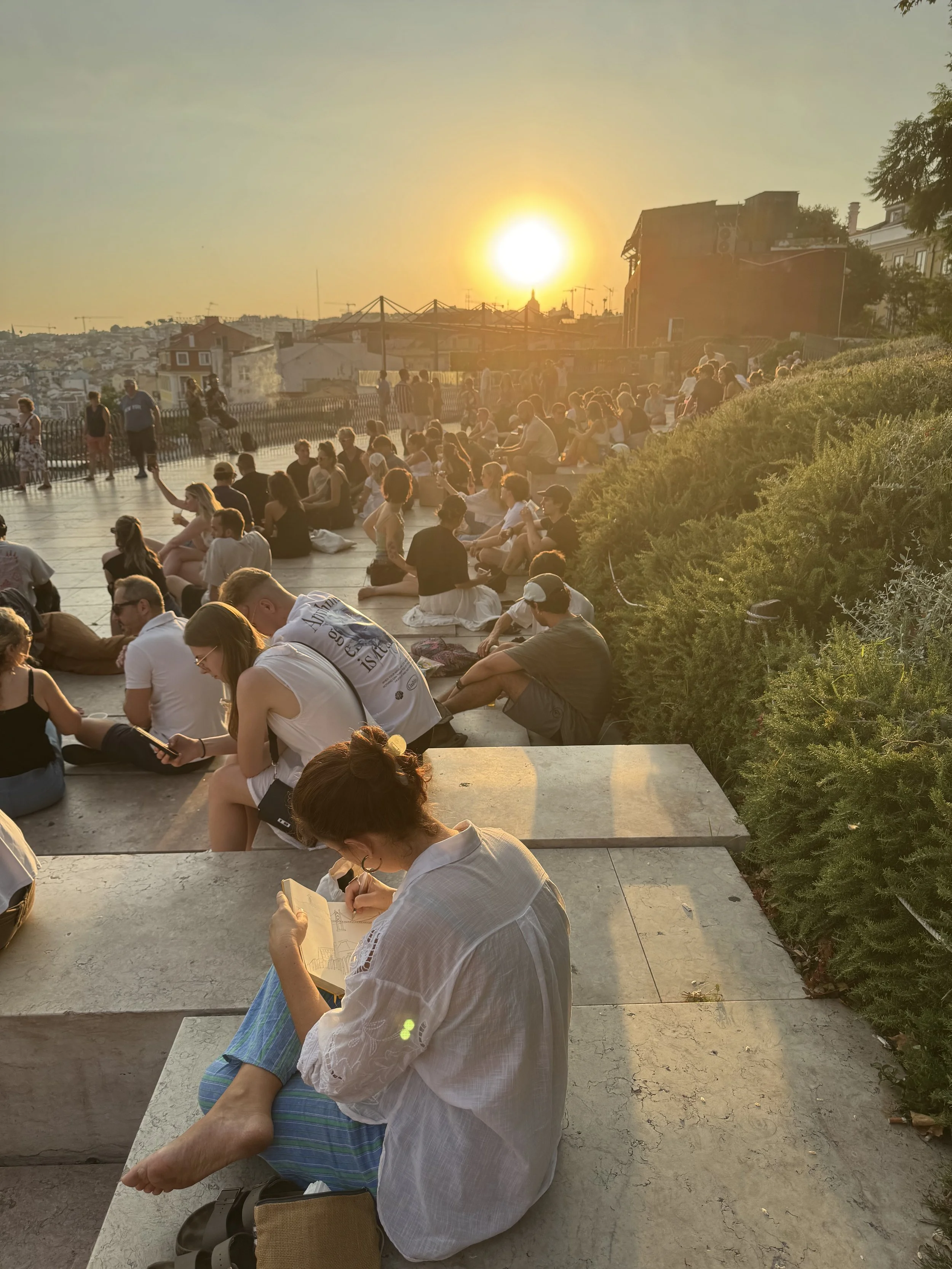 Grupo de personas sentadas en un mirador de Lisboa al atardecer, algunas conversando y otras leyendo o escribiendo, con la ciudad de fondo y el sol poniéndose en el horizonte.