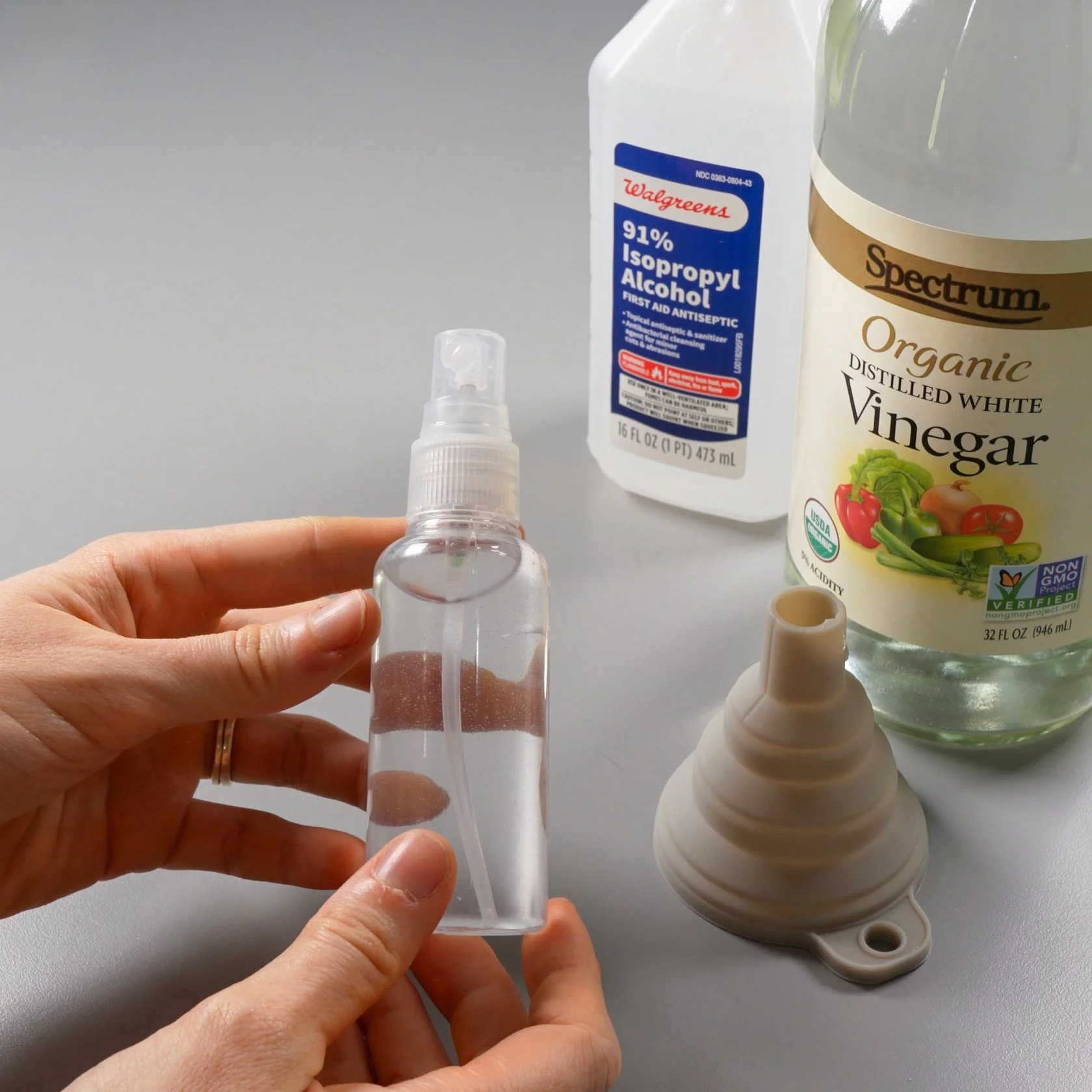 Hands holding a spray bottle of isopropyl alcohol, next to a funnel and a bottle of white vinegar