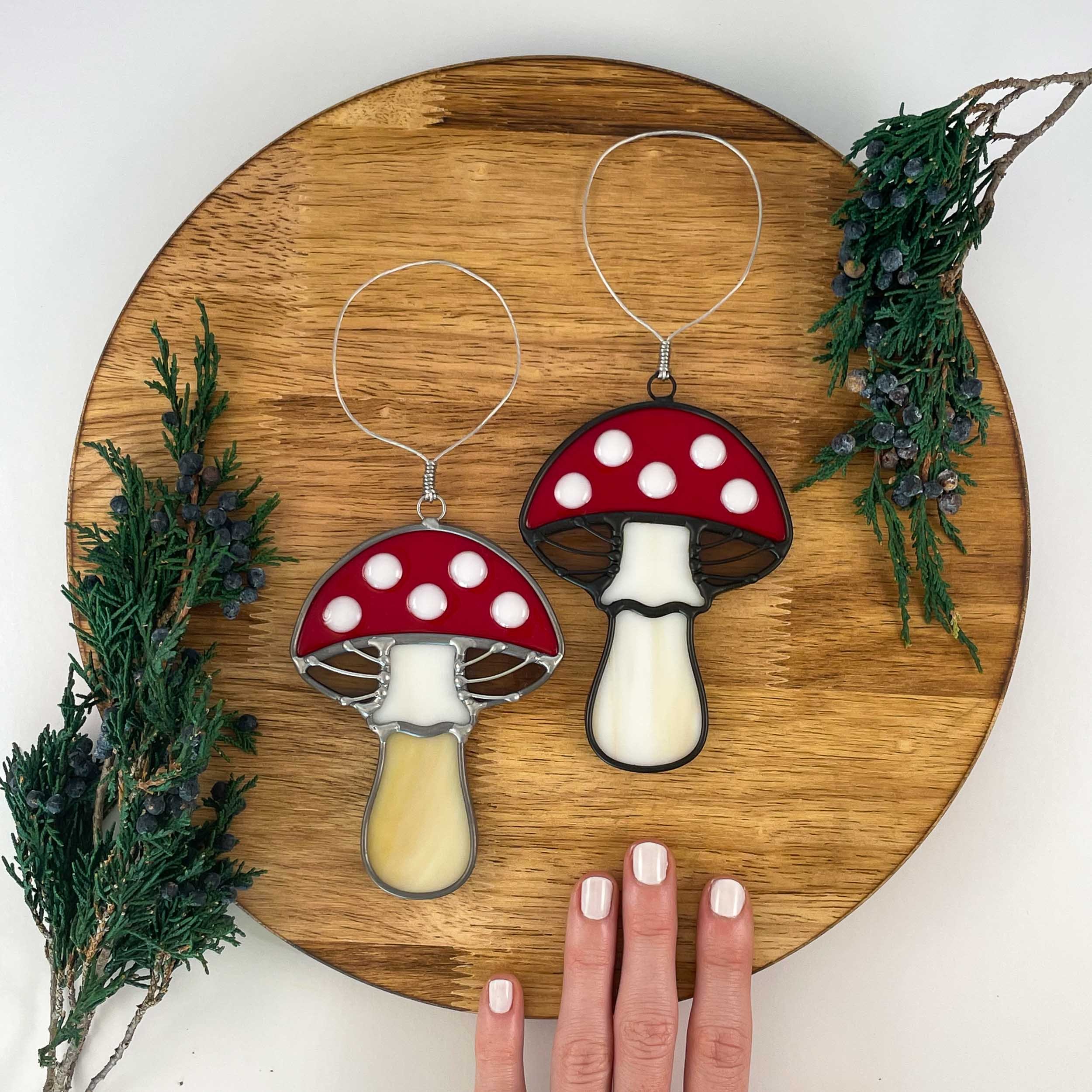 Two stained glass toadstool mushroom ornaments with white-spotted red caps arranged on a wooden board next to Juniper springs