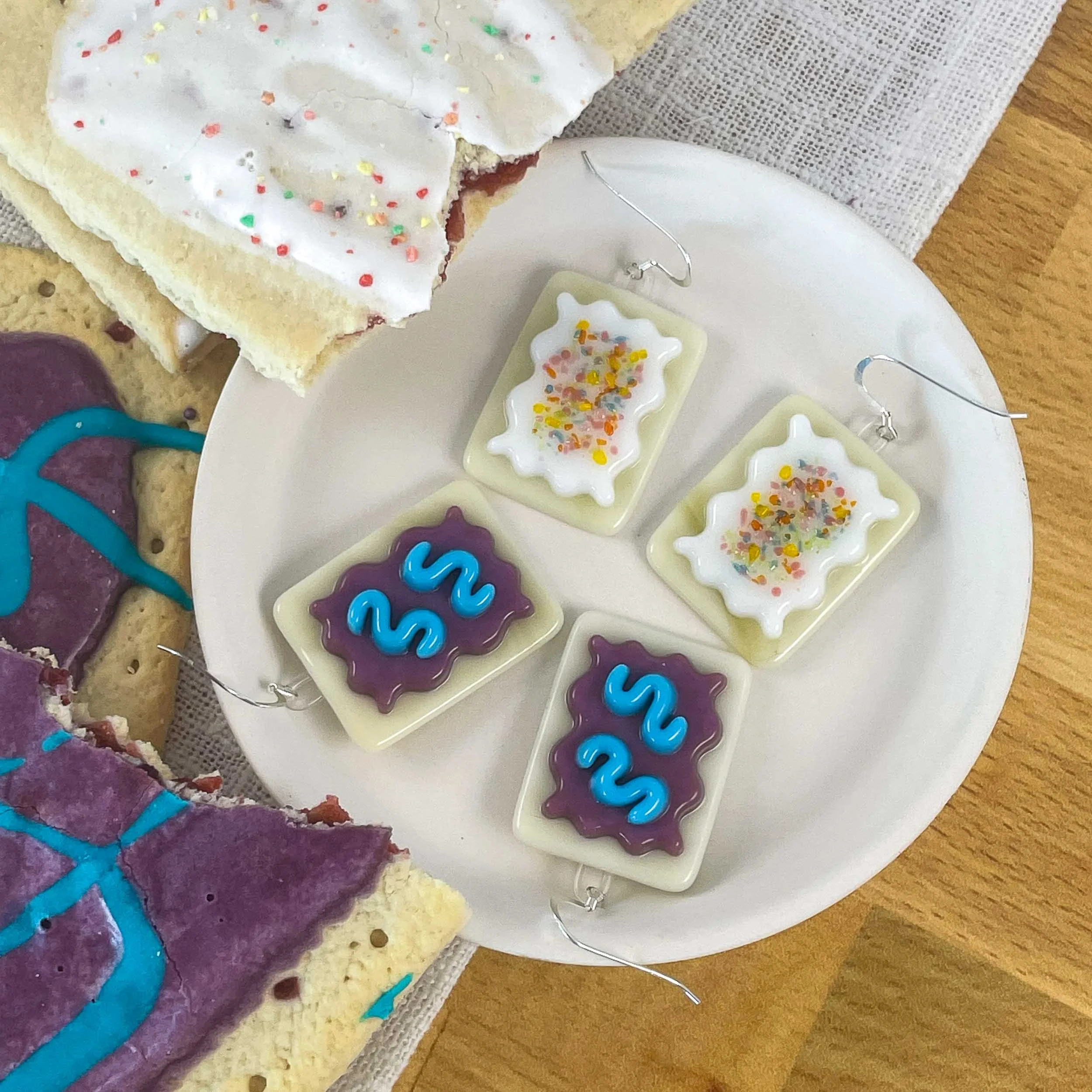 Two pairs of fused glass Pop-Tart earrings on a plate next to actual Pop-Tarts
