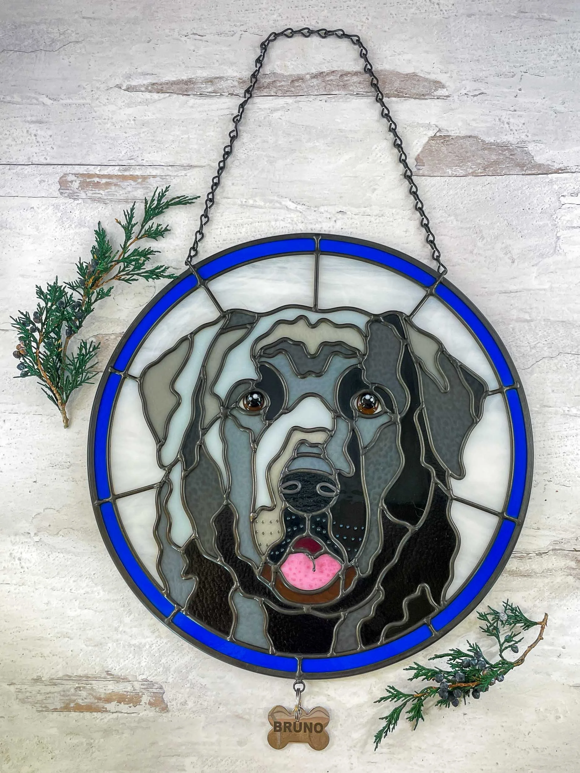 A circular stained glass pet portrait panel of a black Labrador dog with a bone-shaped nametag reading "Bruno" dangling from its lower border