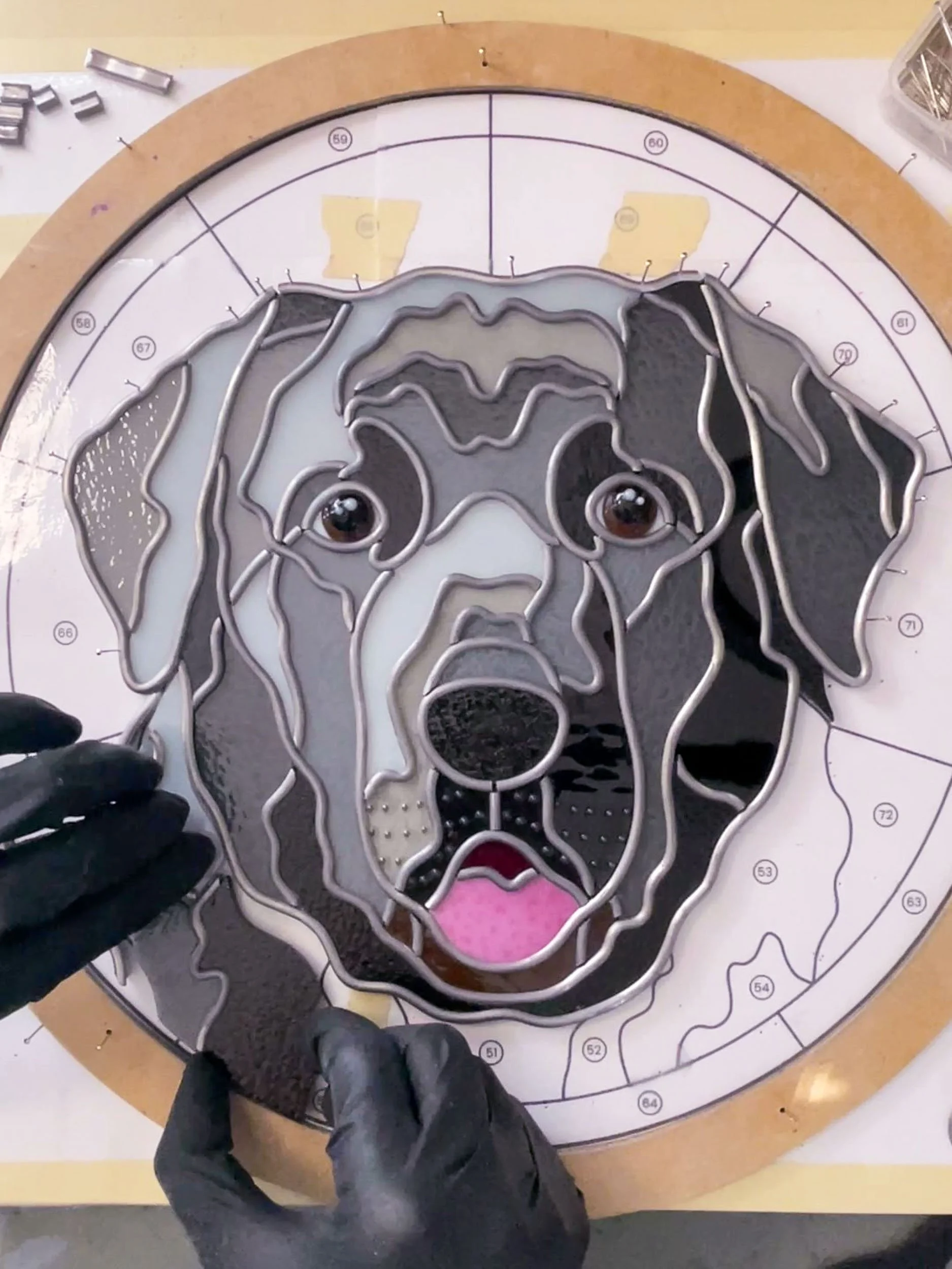 A pair of gloved hands placing together leaded pieces of a stained glass dog pet portrait