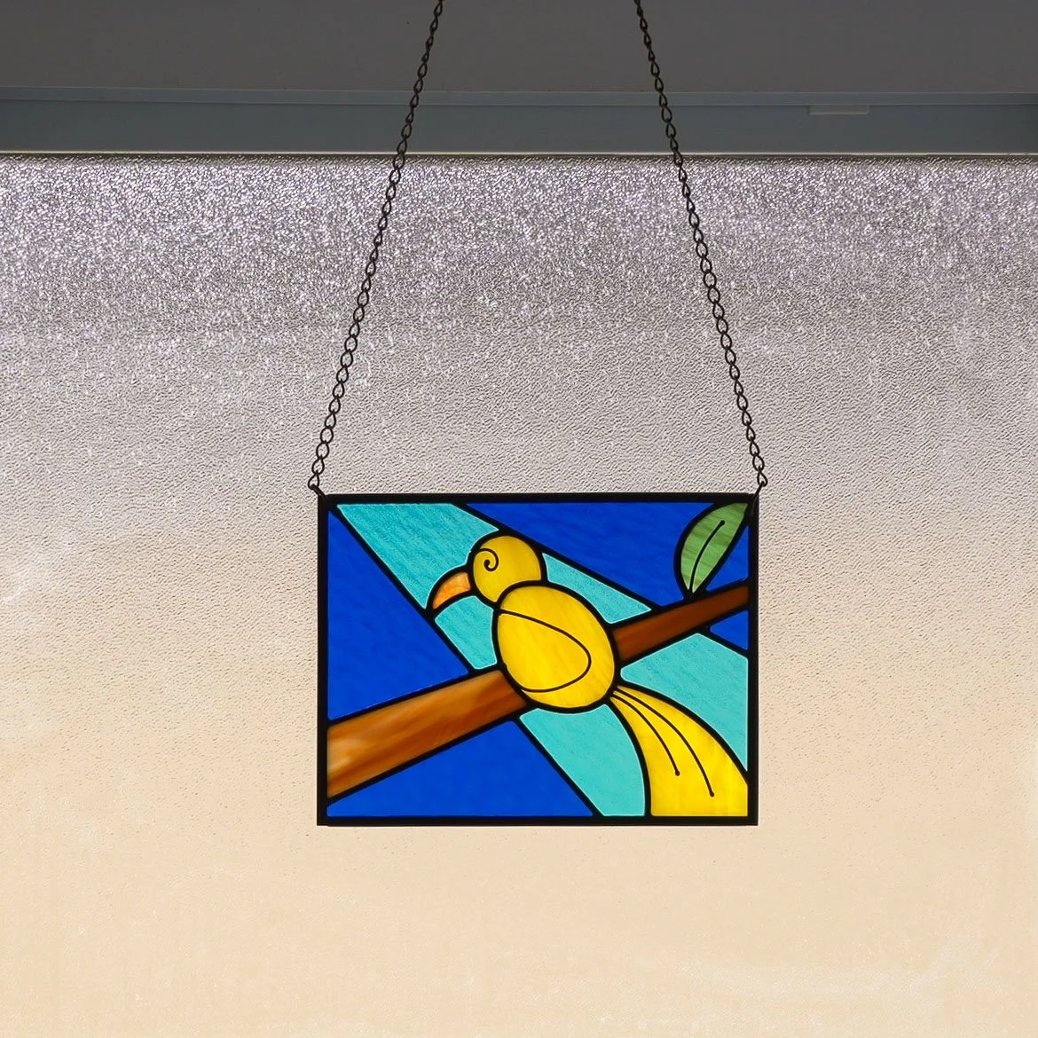 A colorful stained glass panel depicting a yellow canary sitting on a tree branch, hanging in a sunlit window
