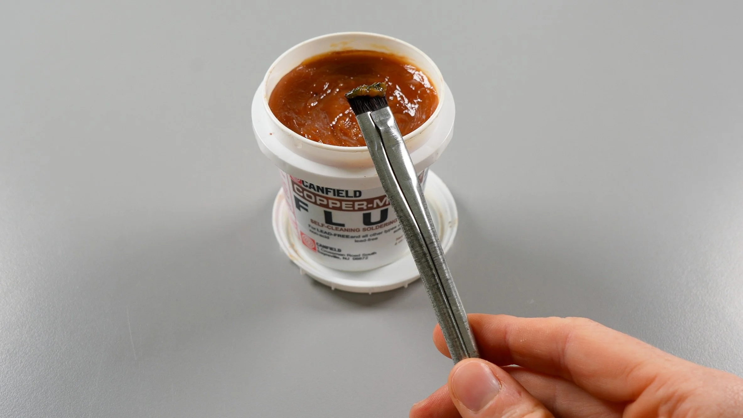 A hand holding a flux brush in front of a container of stained glass paste flux