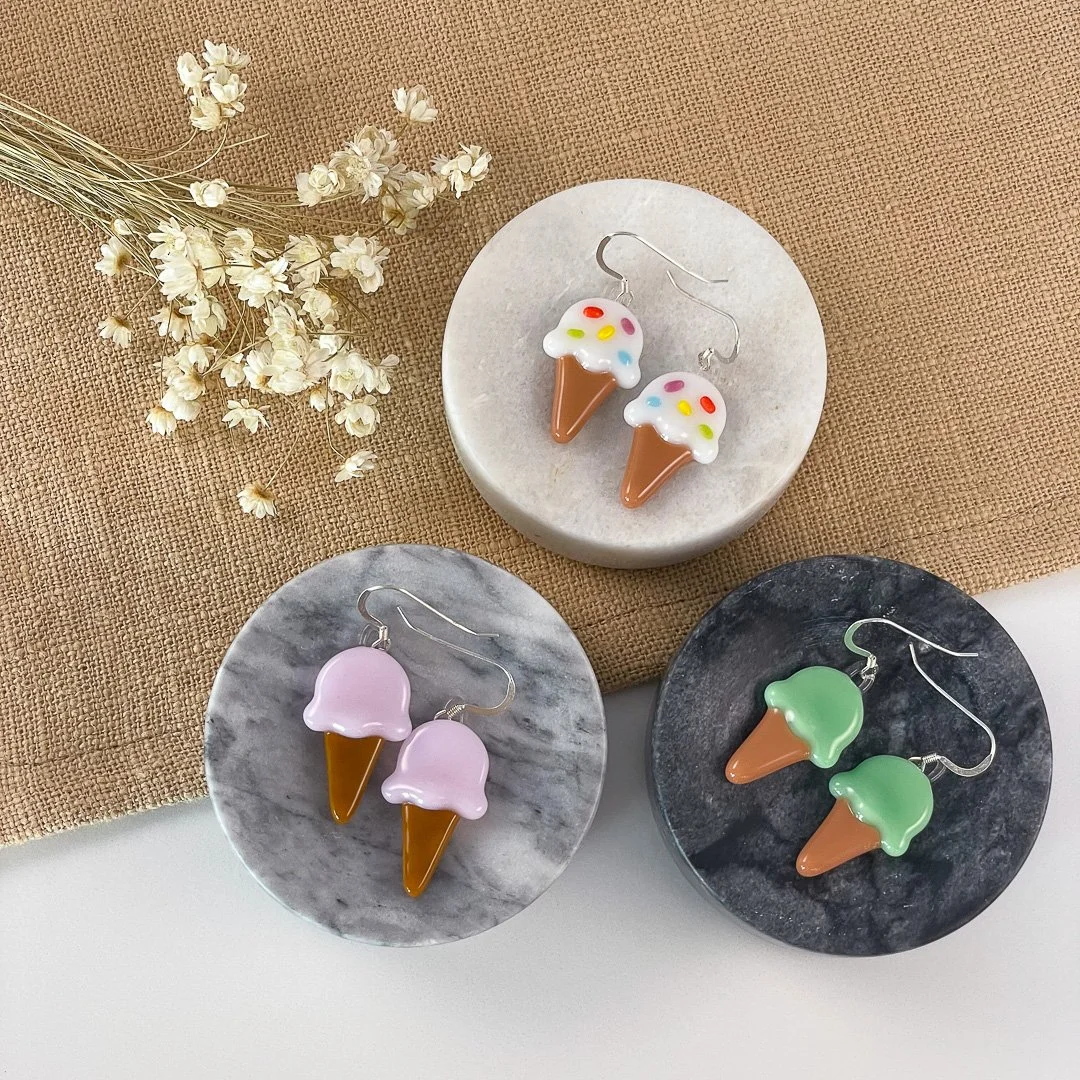 Pairs of various-colored fused glass ice cream cone earrings arranged on marble dishes