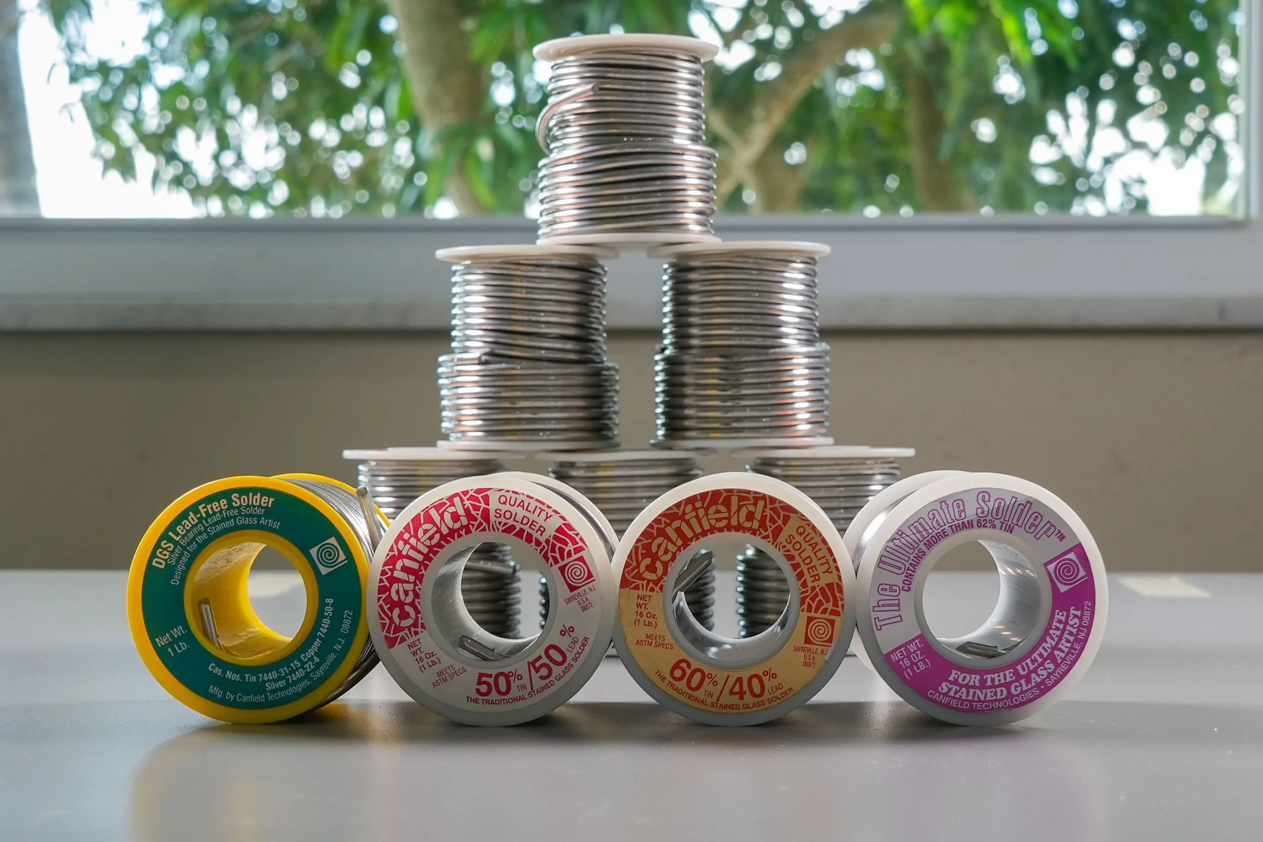 Spools of various types of stained glass solder laid in front of a pyramid of stacked solder spools