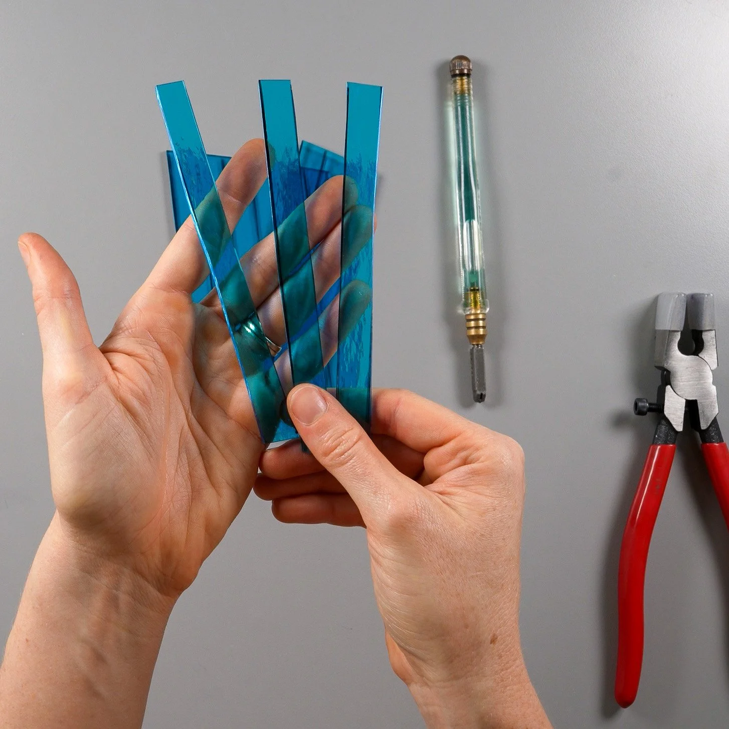 Hands holding several long, thin strips of stained glass next to a glass cutter and running pliers