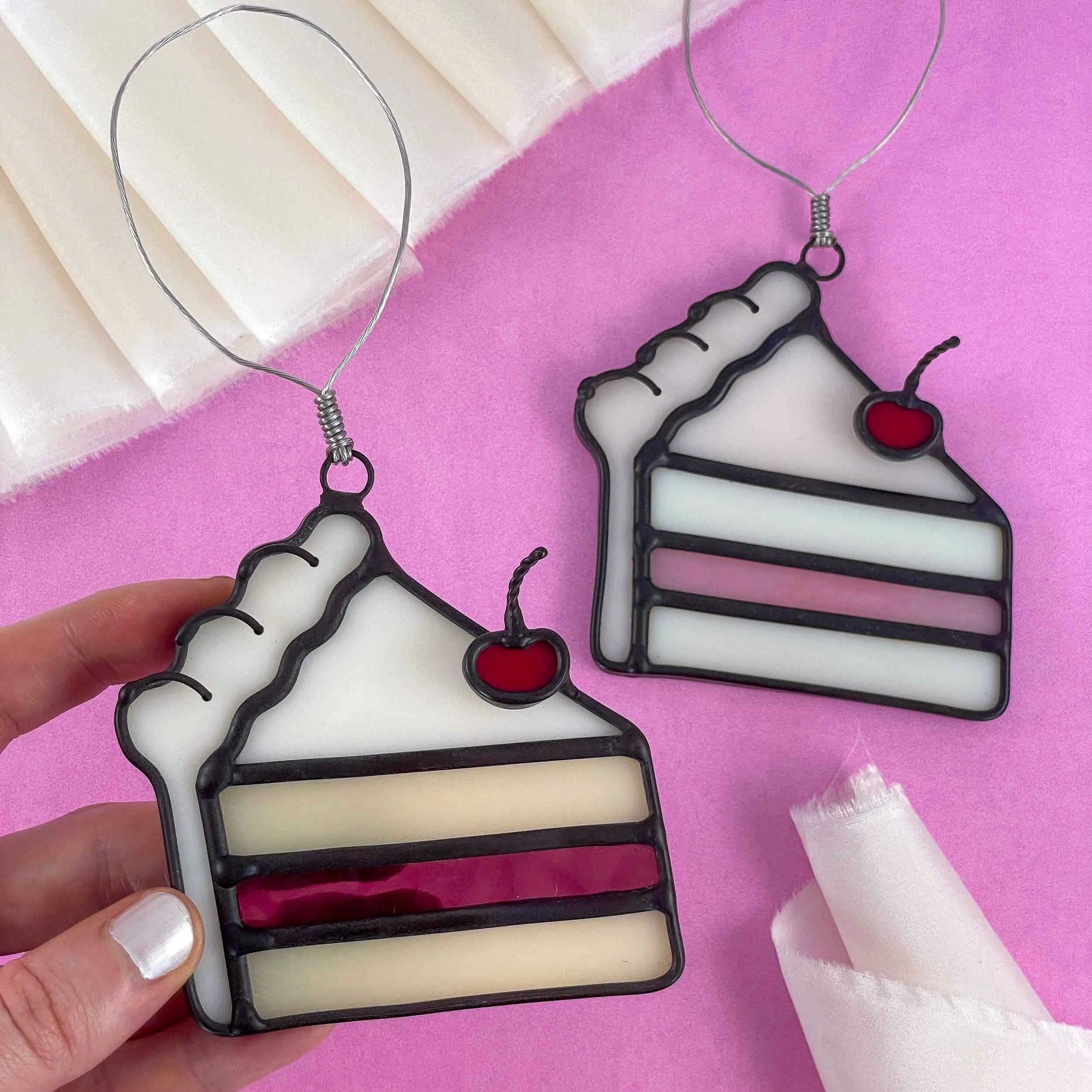 Two different colored stained glass cake slice ornaments arranged on a bright pink background