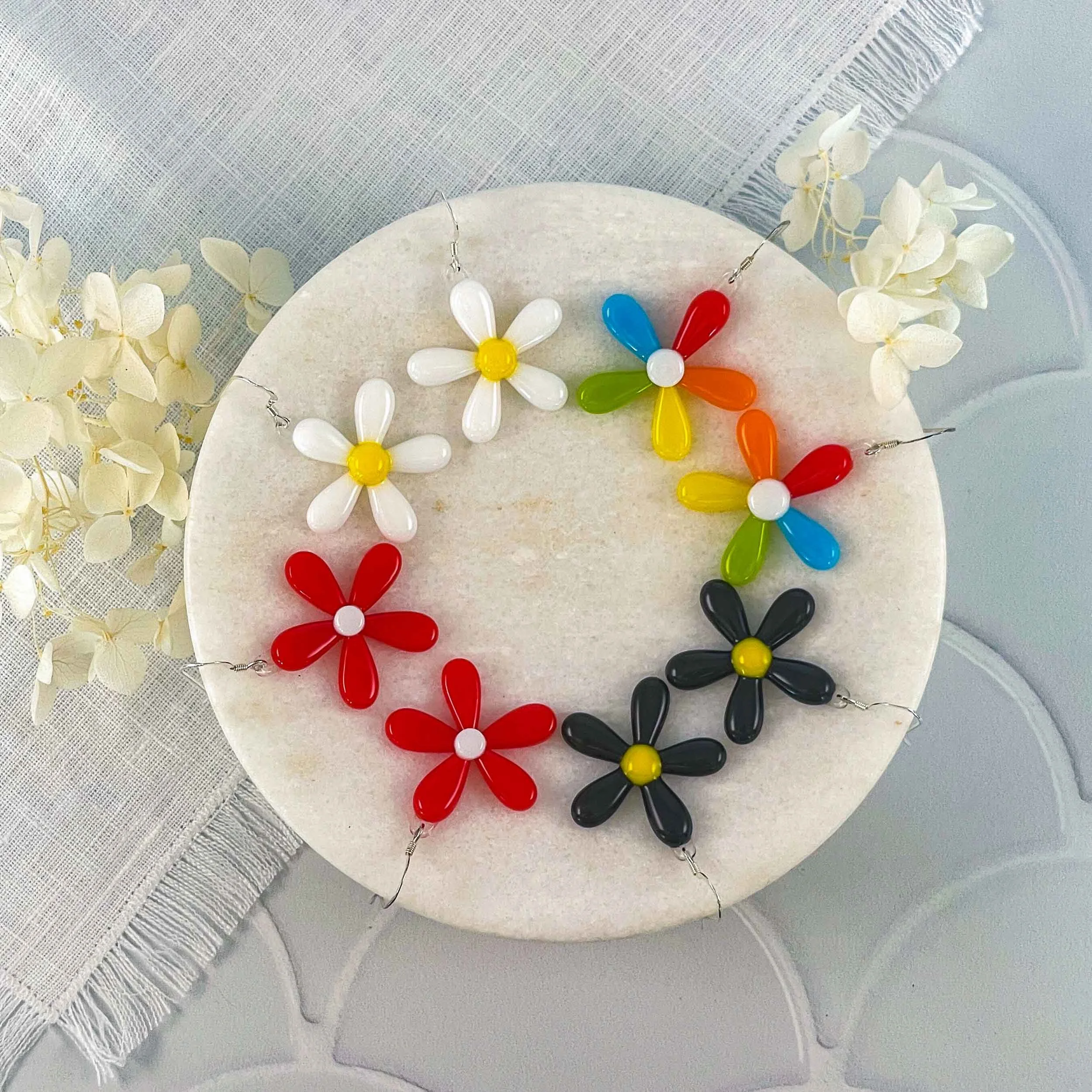 Various colored fused glass daisy earrings arranged around the edge of a circular marble dish