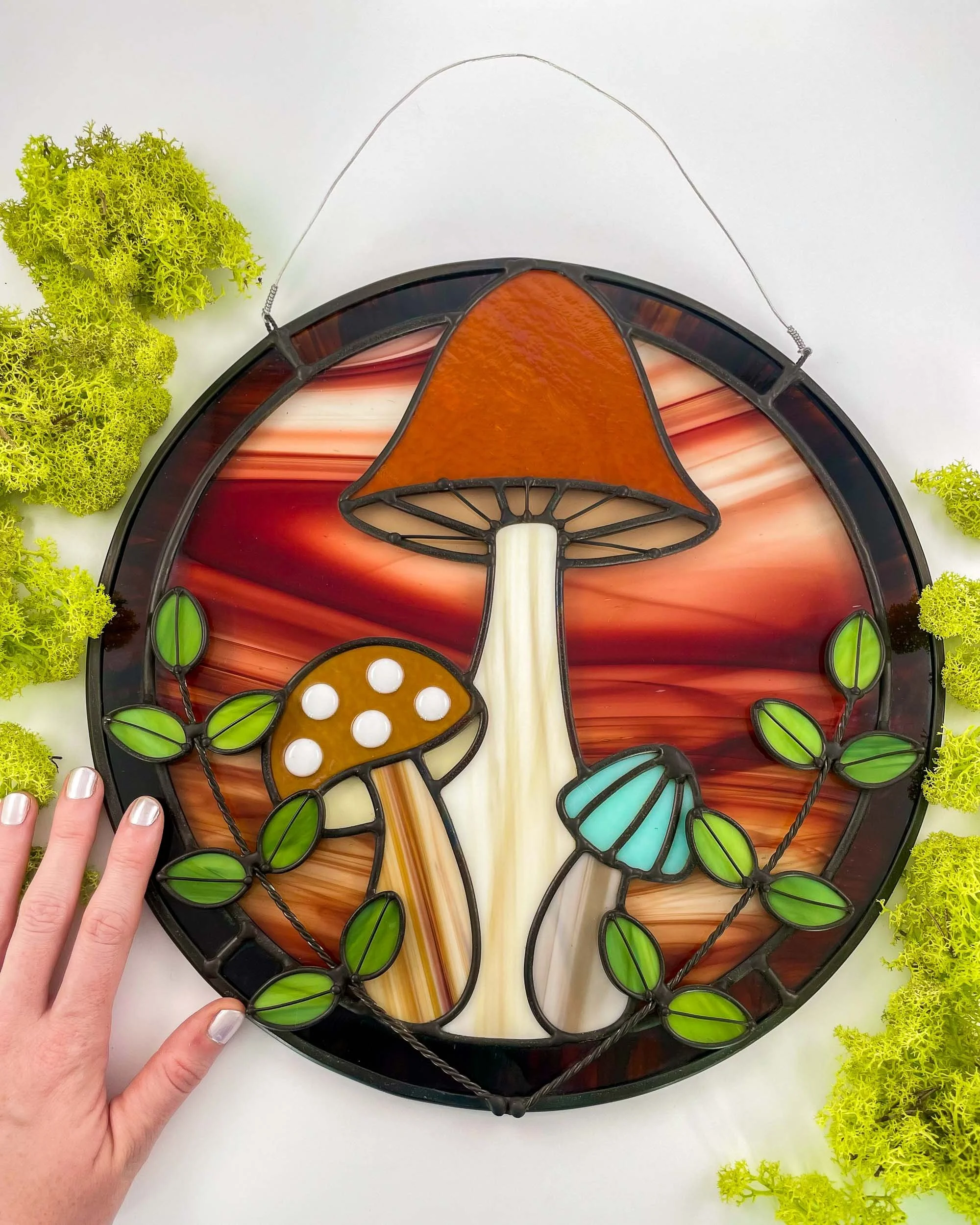 A vibrantly colored stained glass panel depicting a trio of different sized mushrooms, laid next to green moss