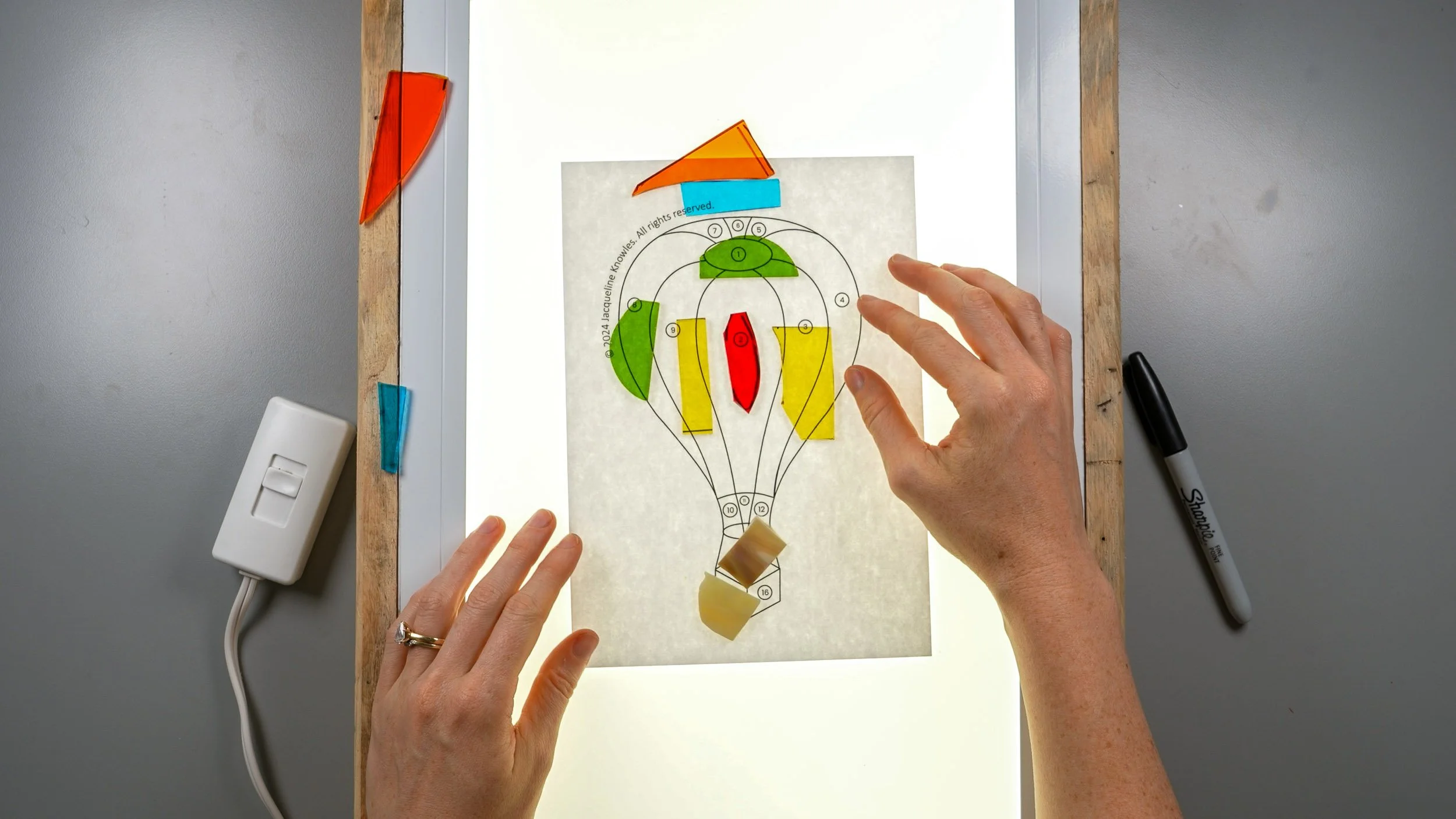 Colorful stained glass scraps being placed atop a hot air balloon pattern over a lightboard