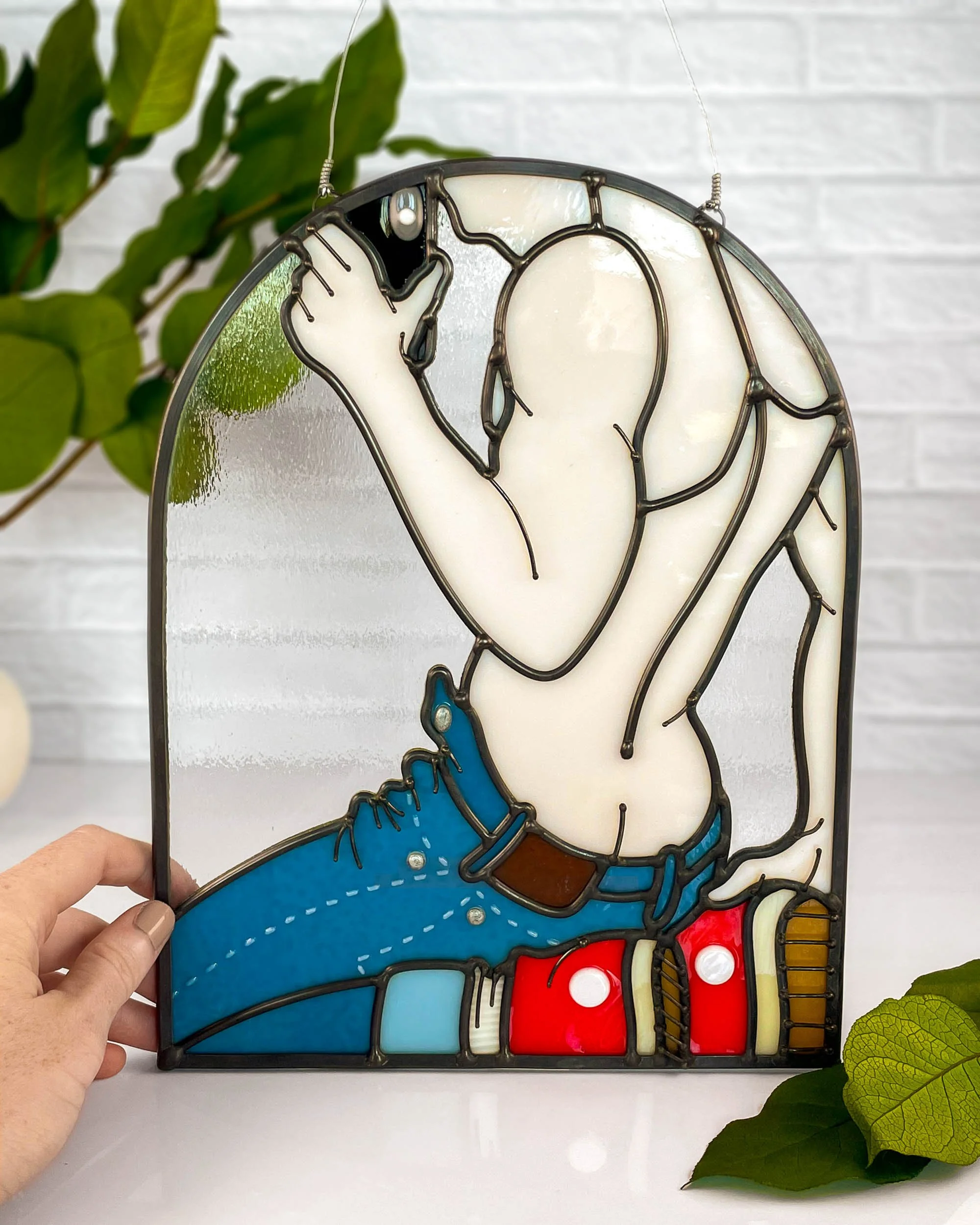 A stained glass panel depicting a shirtless man wearing unzipped blue jeans and posing in a flirtatious way while taking a selfie