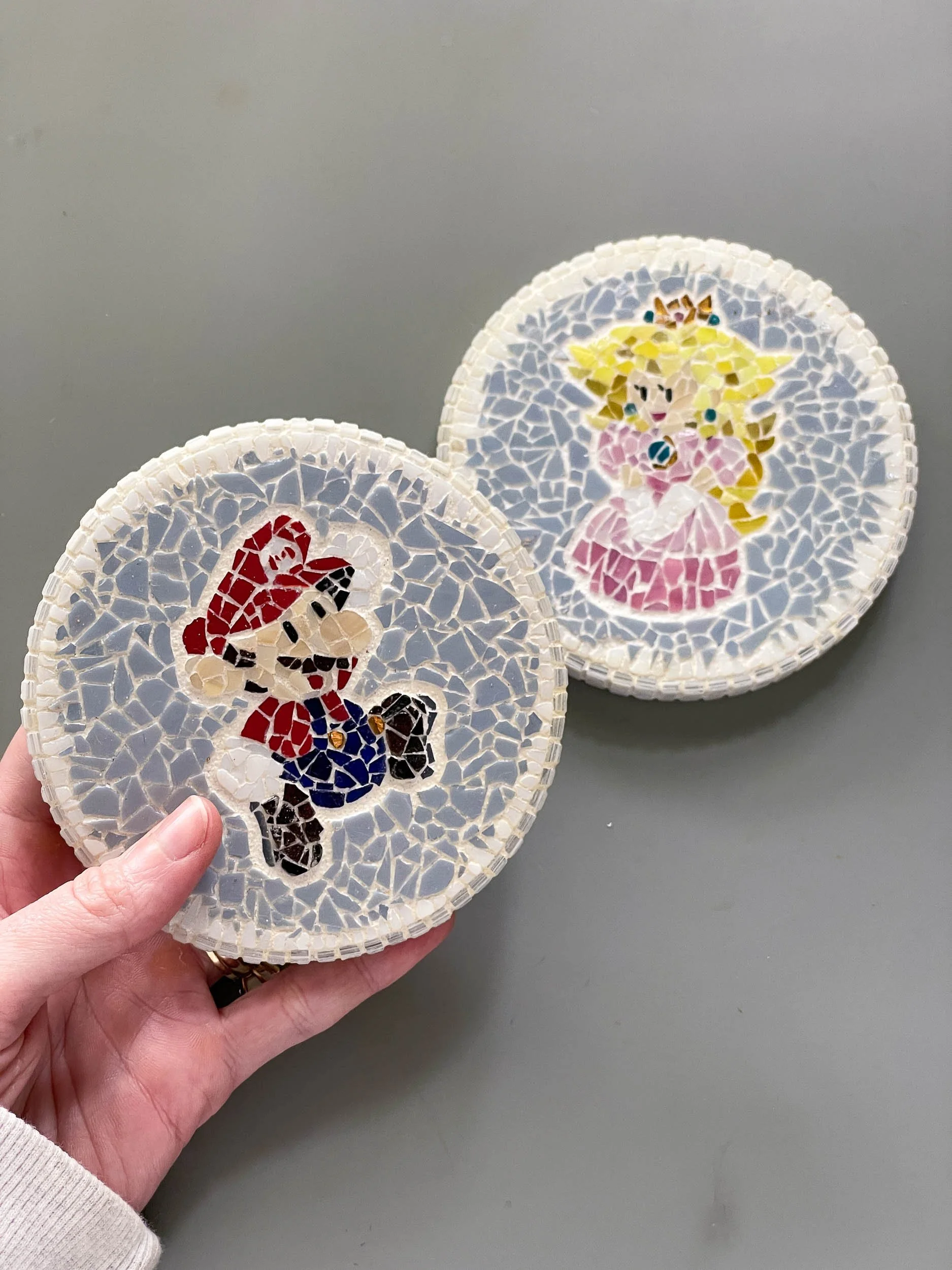 A hand holding Mario-themed stained glass mosaic coasters handmade by Glasswork Pixie