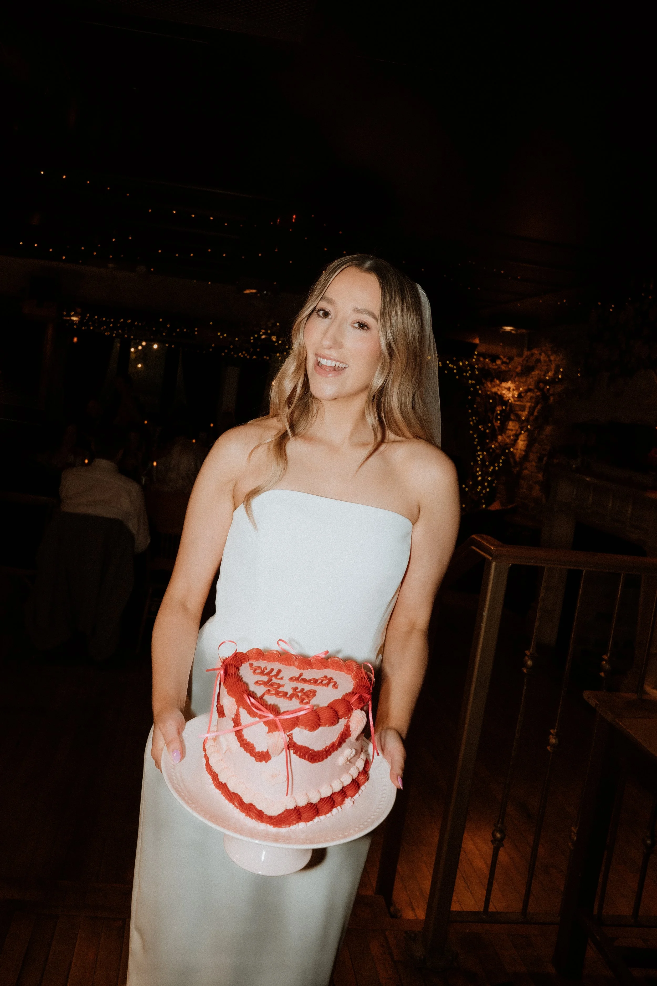 A woman in a white strapless dress holding a pink and red heart-shaped cake with the words 'still death do us part' written on it, likely at a wedding reception.