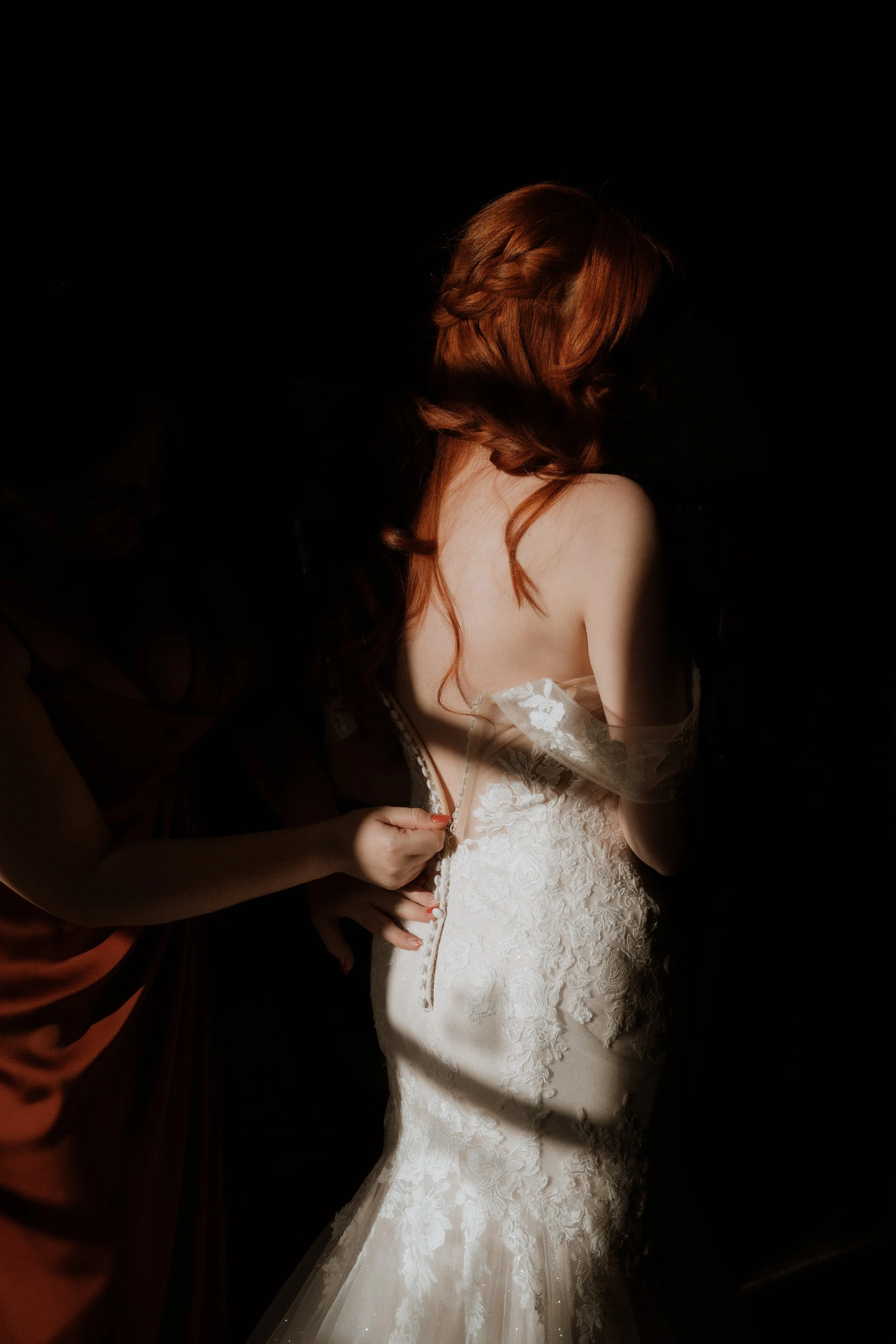 A woman in a wedding dress being buttoned up by another person in a dark setting.