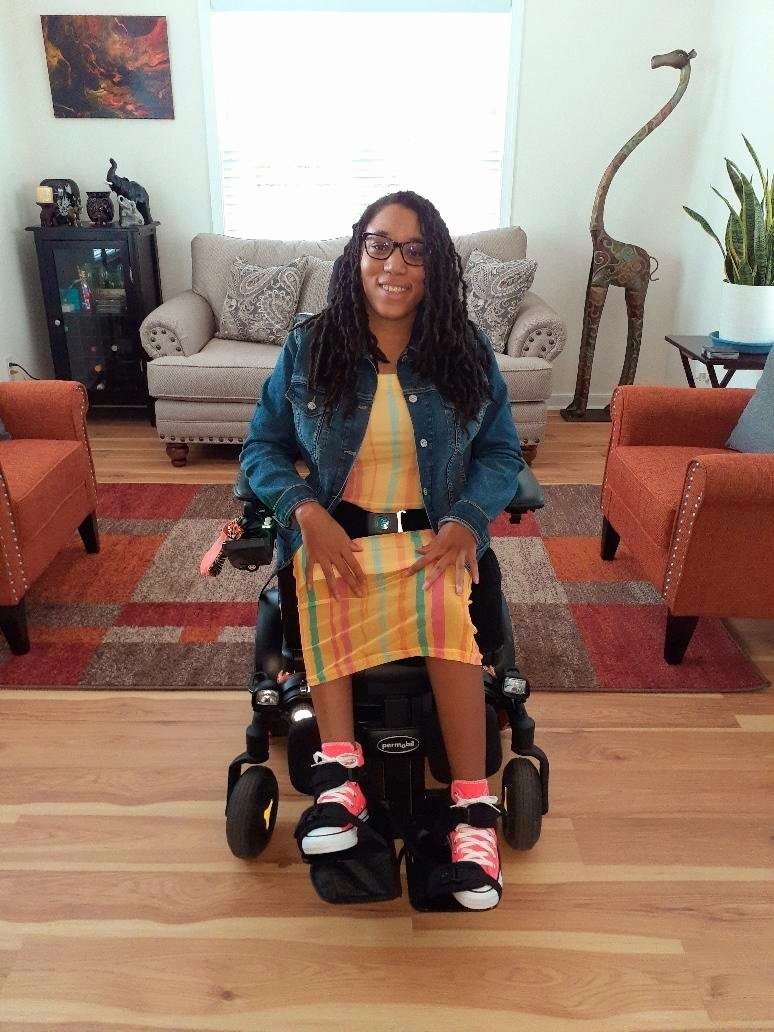  Jessica Johnson in a wheelchair, wearing a yellow dress and a denim jacket on top. Pictured in front of a sofa.  