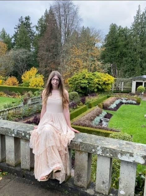  Anah wearing a light pink maxi dress and sitting in front of a garden and trees.  