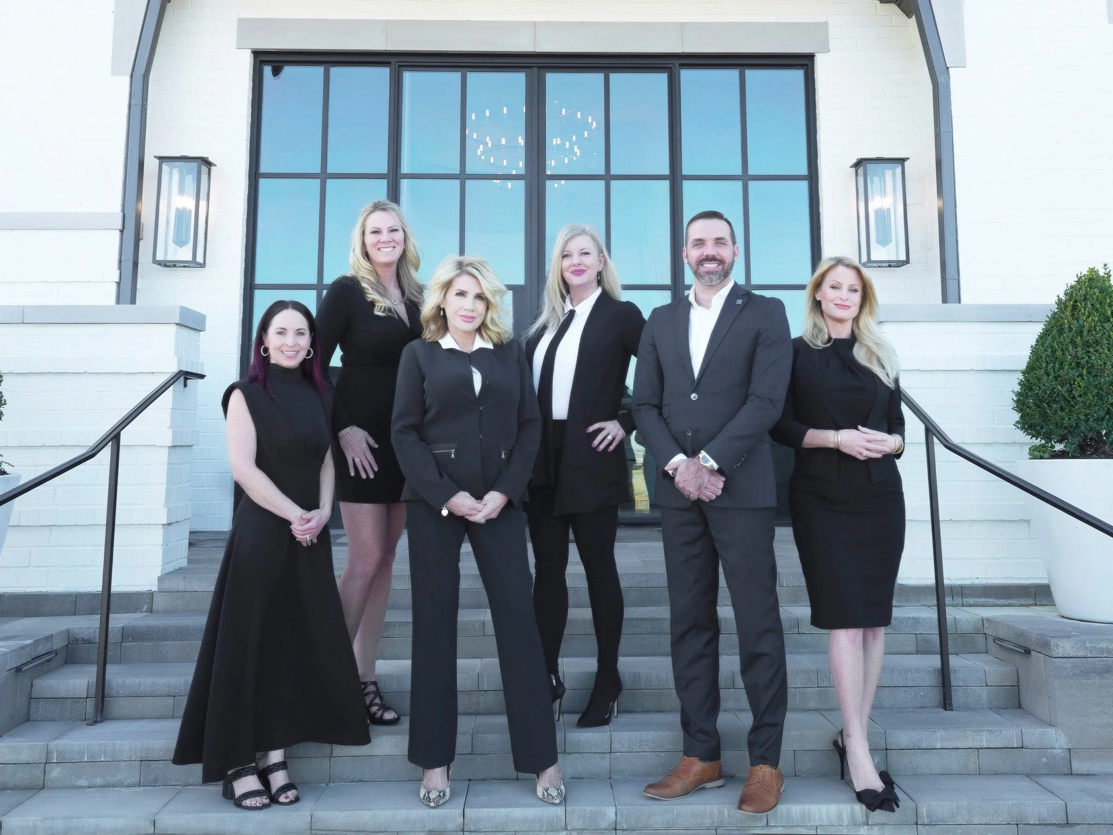 Group of seven professionally dressed people standing on the steps in front of a large glass window of a building.