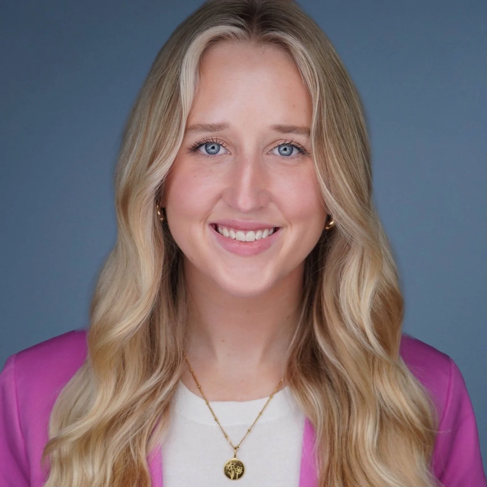 A smiling woman with long wavy blonde hair, blue eyes, wearing a white top, pink blazer, gold necklace with a tree pendant, and small gold earrings against a gray background.