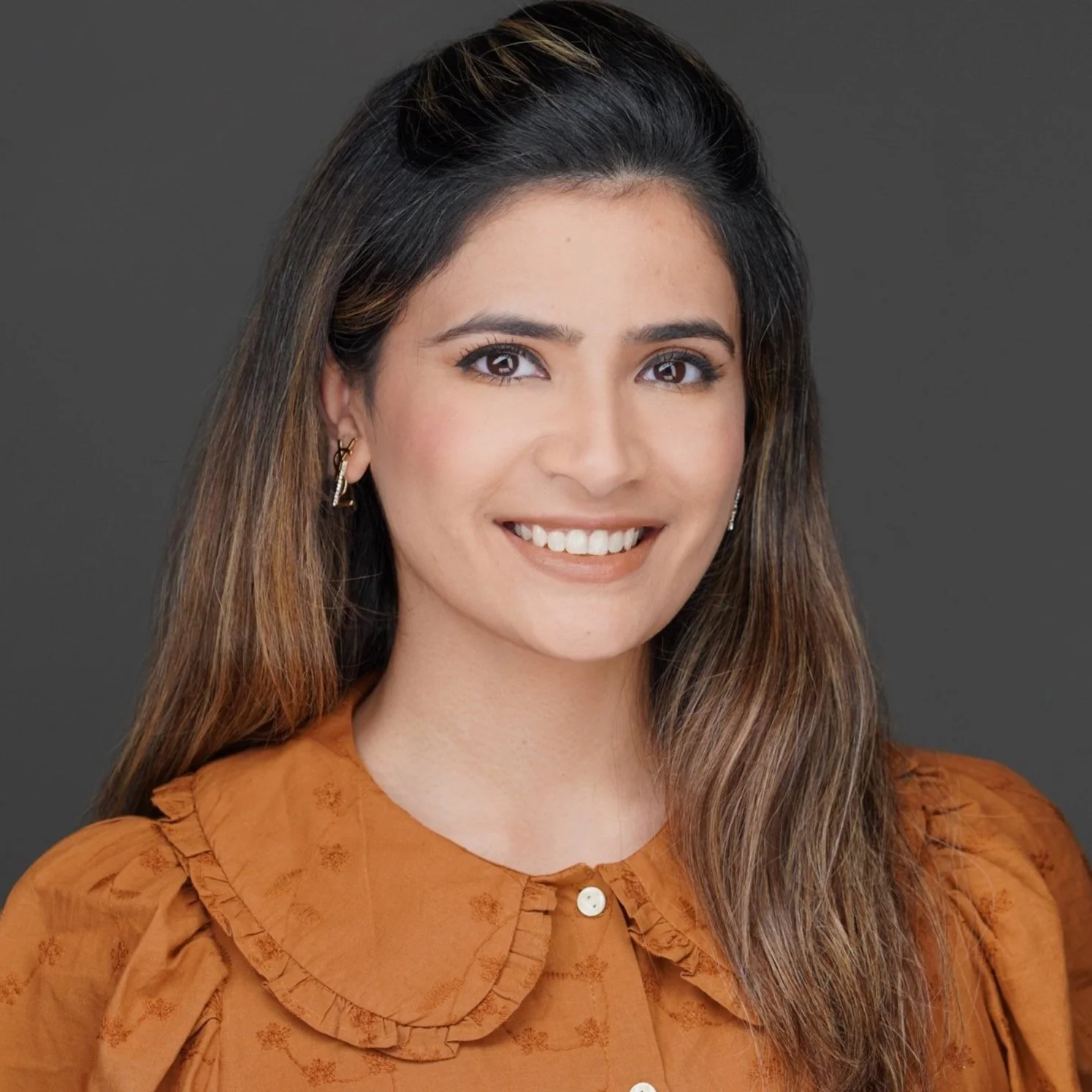 A smiling woman with long brown hair facing forward against a gray background, wearing an orange blouse with a Peter Pan collar and small earrings.