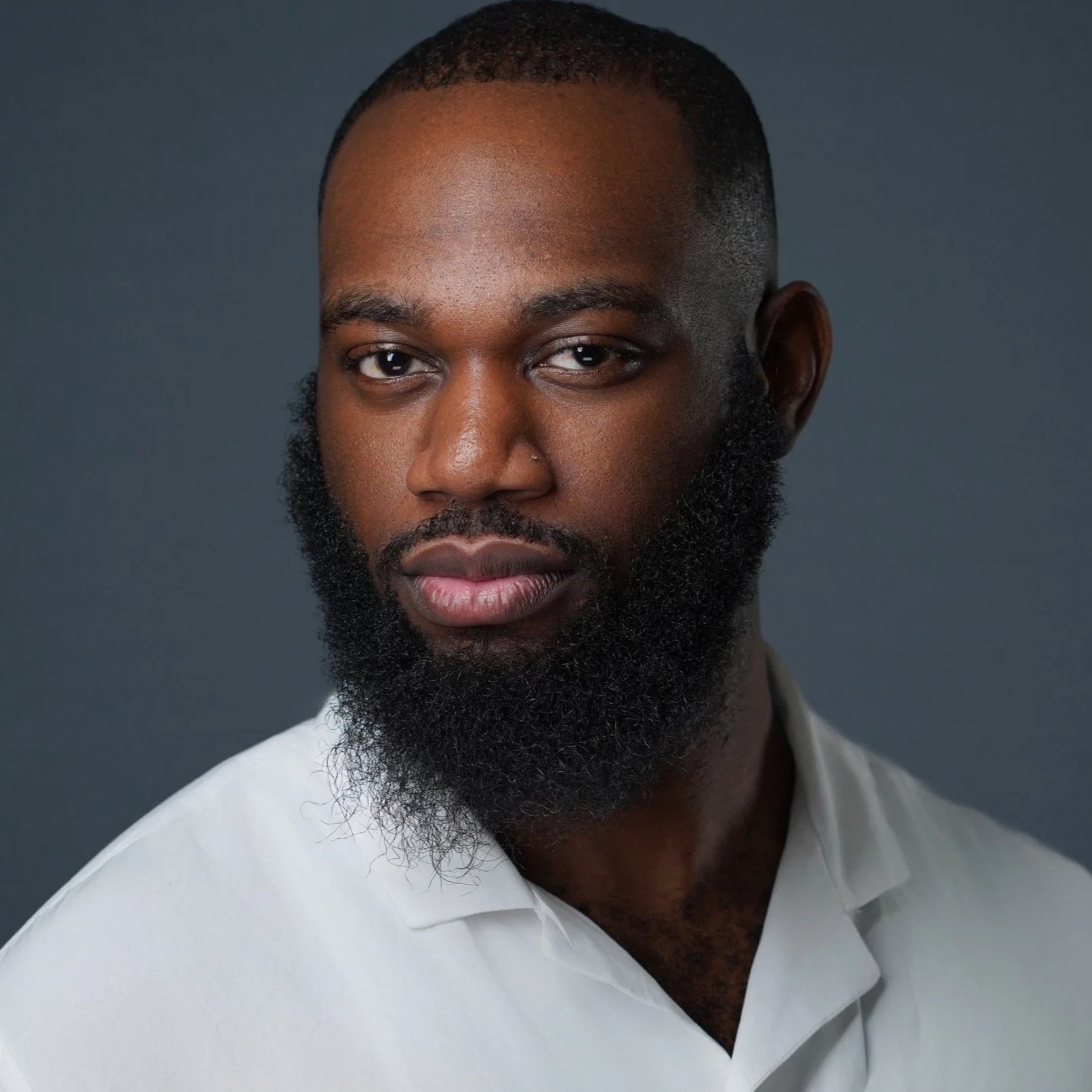 A young man with dark skin, a beard, and short hair, wearing a white shirt, posing against a dark background.